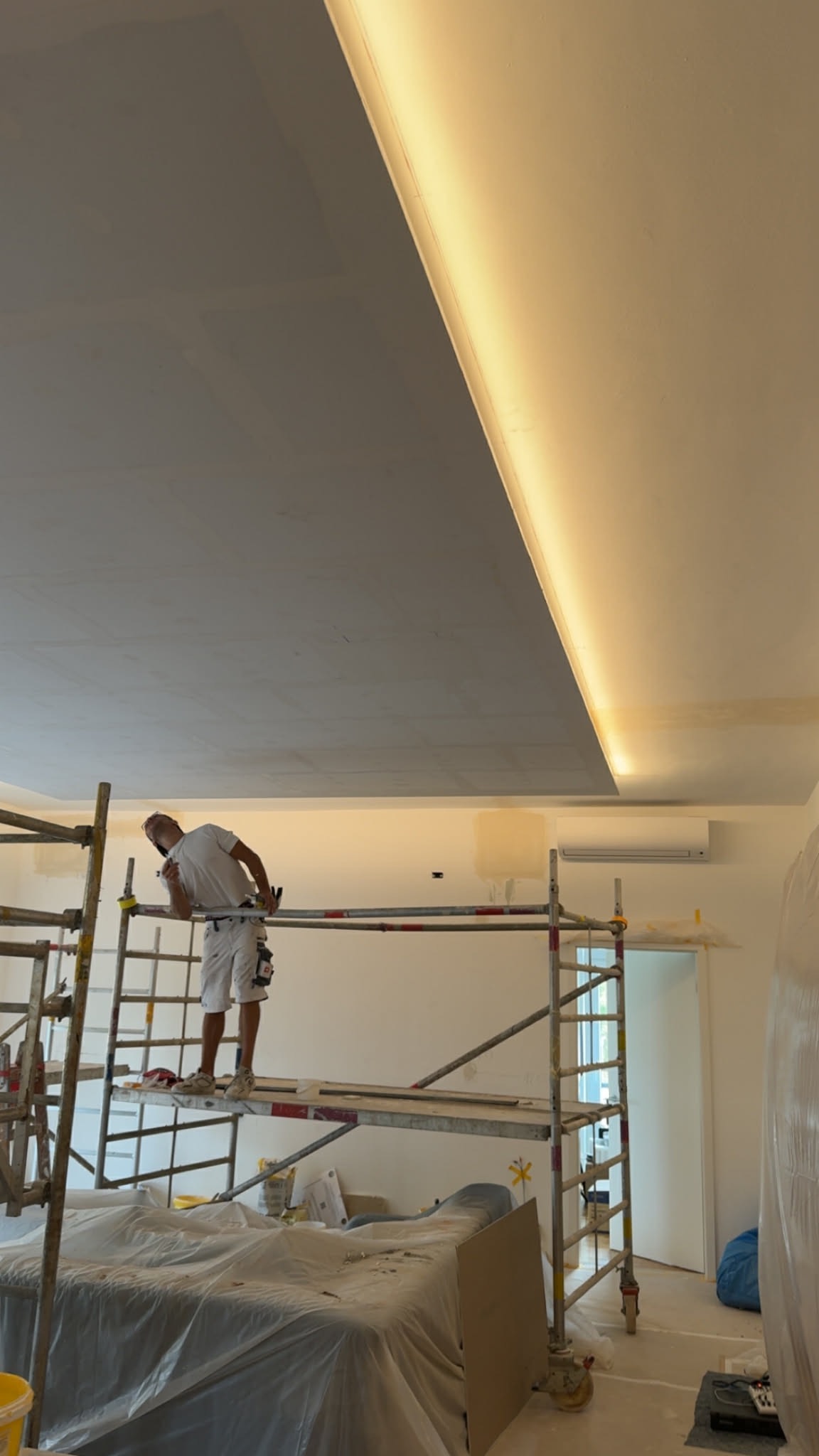 Malarz na rusztowaniu maluje sufit z oświetleniem LED. Widoczne zabezpieczone meble i narzędzia. Praca w toku, remont mieszkania.