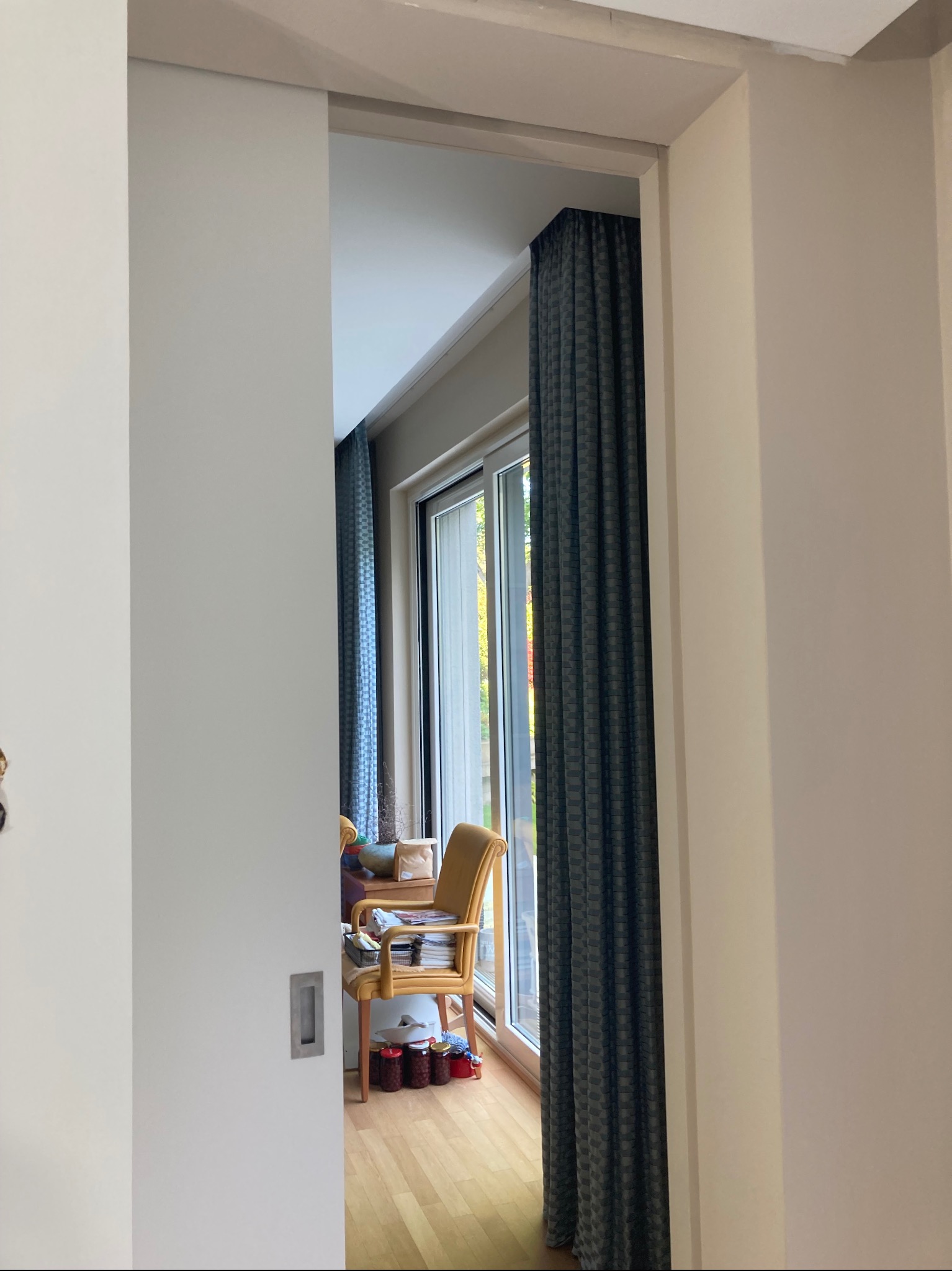 Wnętrze z przesuwnymi drzwiami w kolorze białym, odsłaniające pokój z oknem, zasłonami i krzesłem. Widoczne detale wykończenia ścian i podłogi.