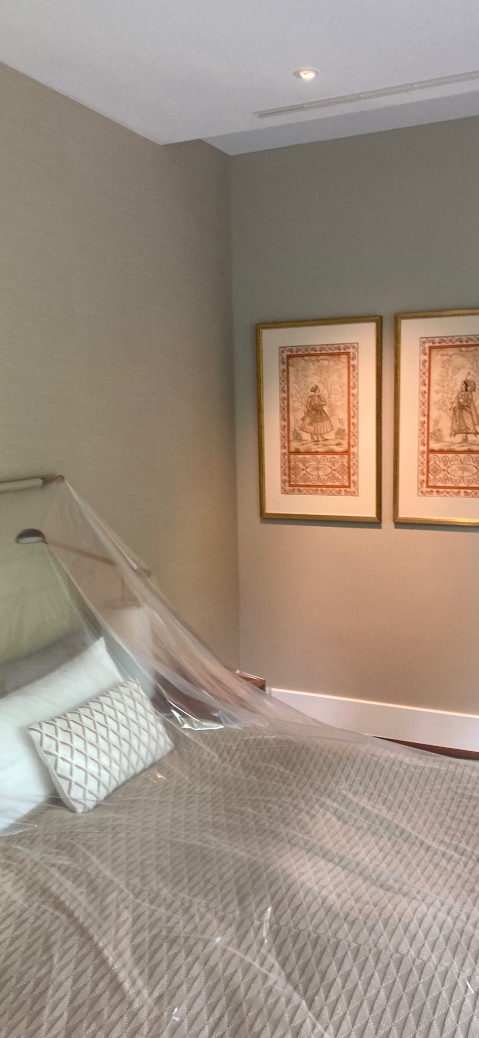 Sypialnia z łóżkiem przykrytym folią ochronną, przygotowana do prac malarskich. Na ścianie wiszą dwa obrazy w złotych ramach. Neutralna kolorystyka wnętrza.