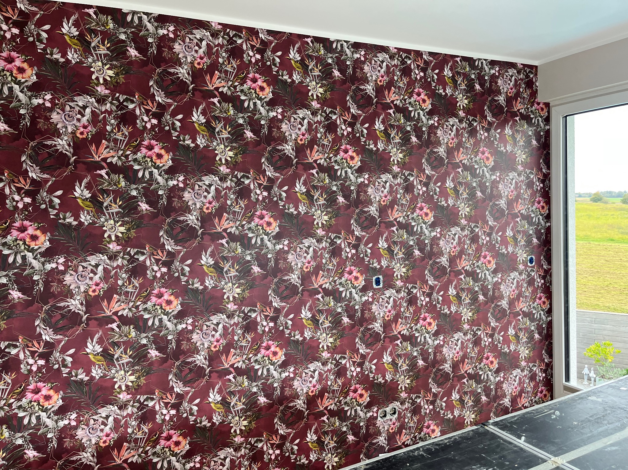 Ściana z bordową tapetą w kwiaty i ptaki, widok z bliska, fragment okna po prawej. Widoczne gniazdka elektryczne. Dekoracyjne wykończenie wnętrza.