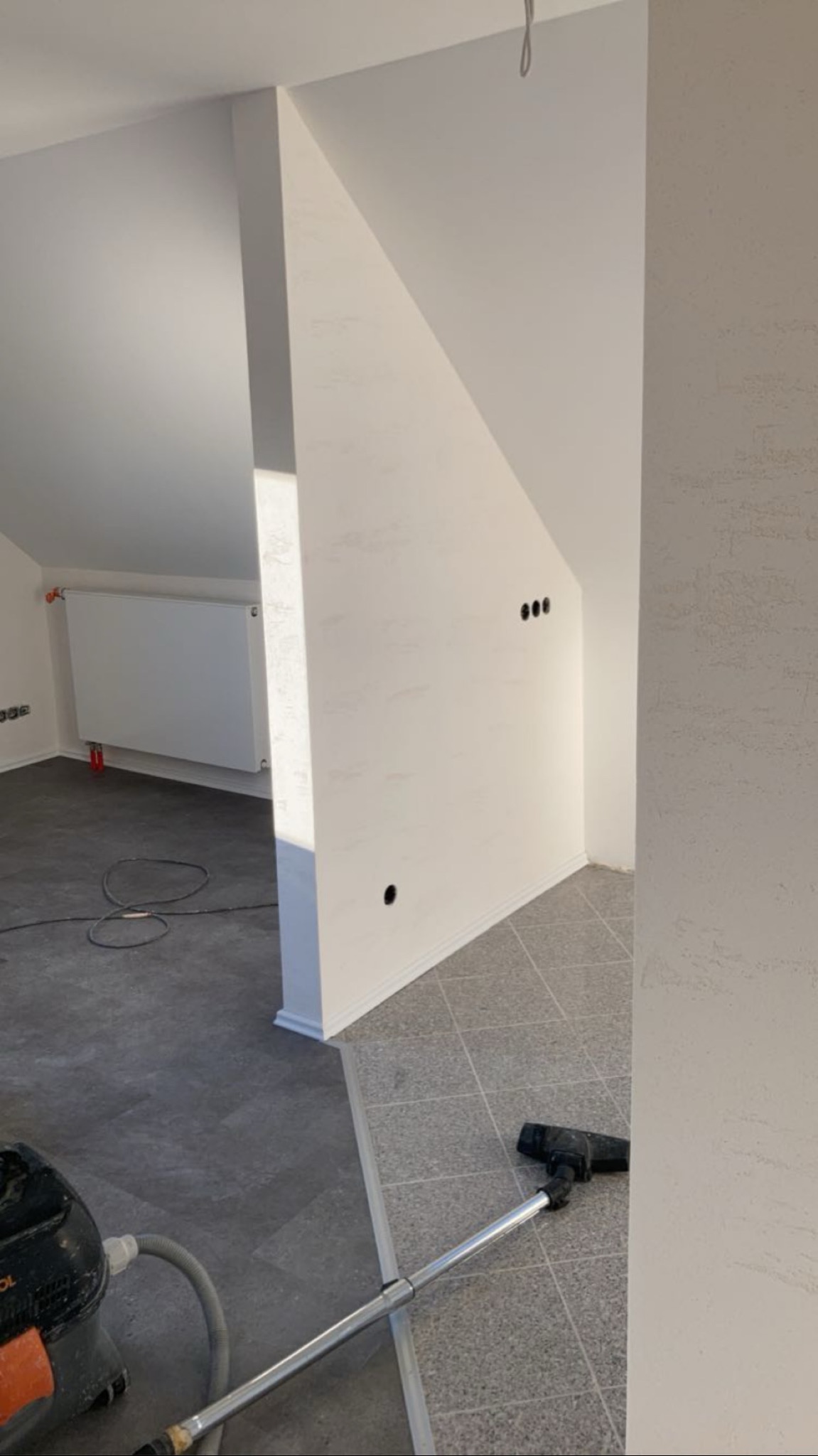 Białe ściany poddasza po malowaniu, widoczne gniazdka elektryczne, grzejnik i odkurzacz. Dwie różne podłogi. Wykończenie wnętrza w jasnych barwach.