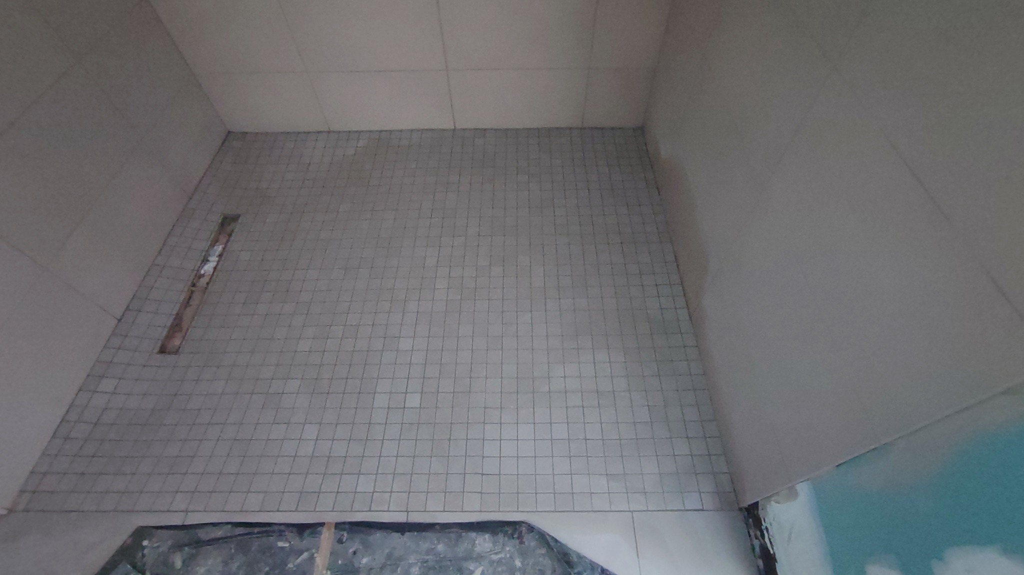 Wykończona podłoga w kabinie prysznicowej z szarych, drobnych płytek. Widoczny odpływ liniowy. Częściowo widoczne zabezpieczenie podłogi folią.