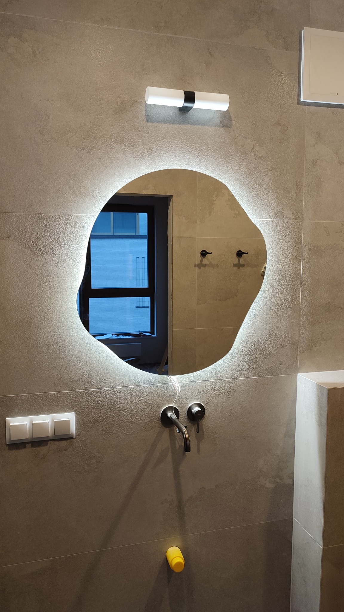 Nowoczesna łazienka z owalnym lustrem LED, szarą płytką imitującą kamień i czarną armaturą. Widoczne okno w odbiciu lustra i biały włącznik światła.