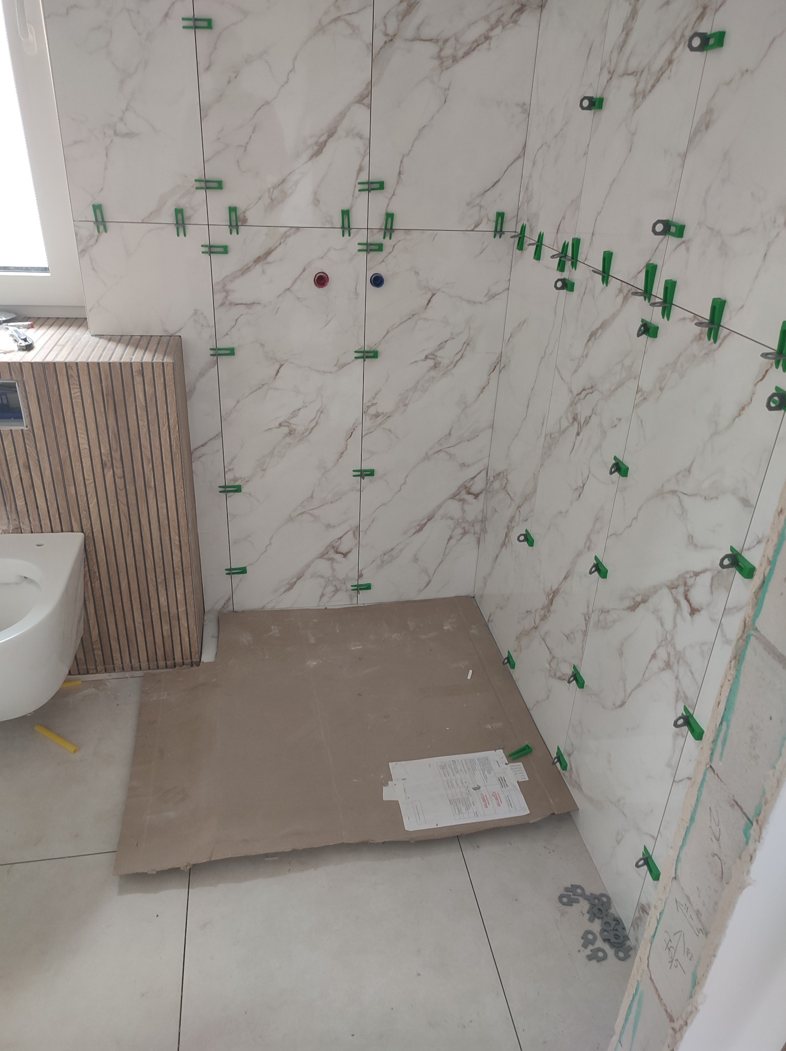 Układanie płytek imitujących marmur w łazience, widoczne zielone klipsy poziomujące, fragment toalety i karton zabezpieczający podłogę. Etap prac wykończeniowych.