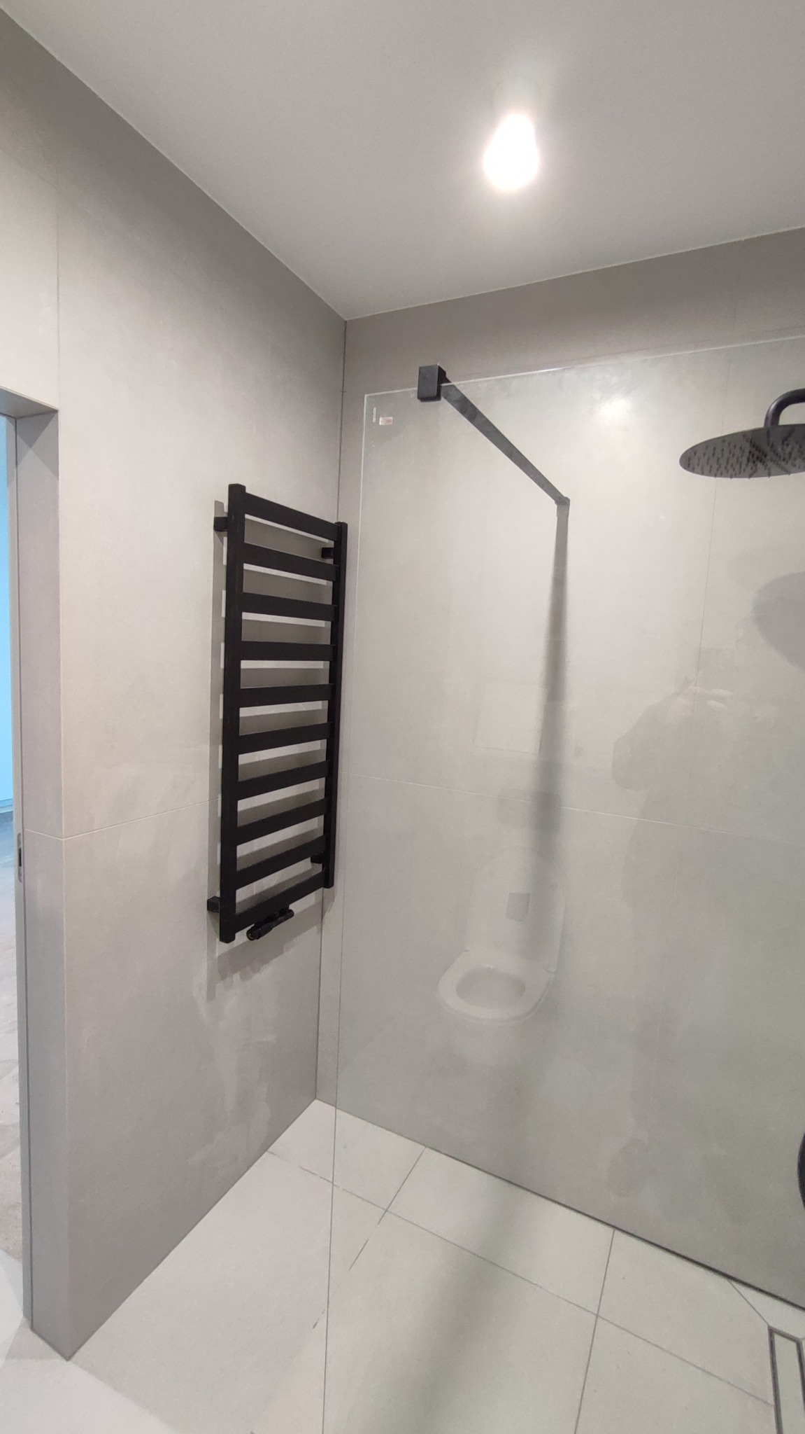 Nowoczesna łazienka z czarnym grzejnikiem drabinkowym i szklaną kabiną prysznicową typu walk-in. Minimalistyczny design, jasne płytki, sufitowe oświetlenie.