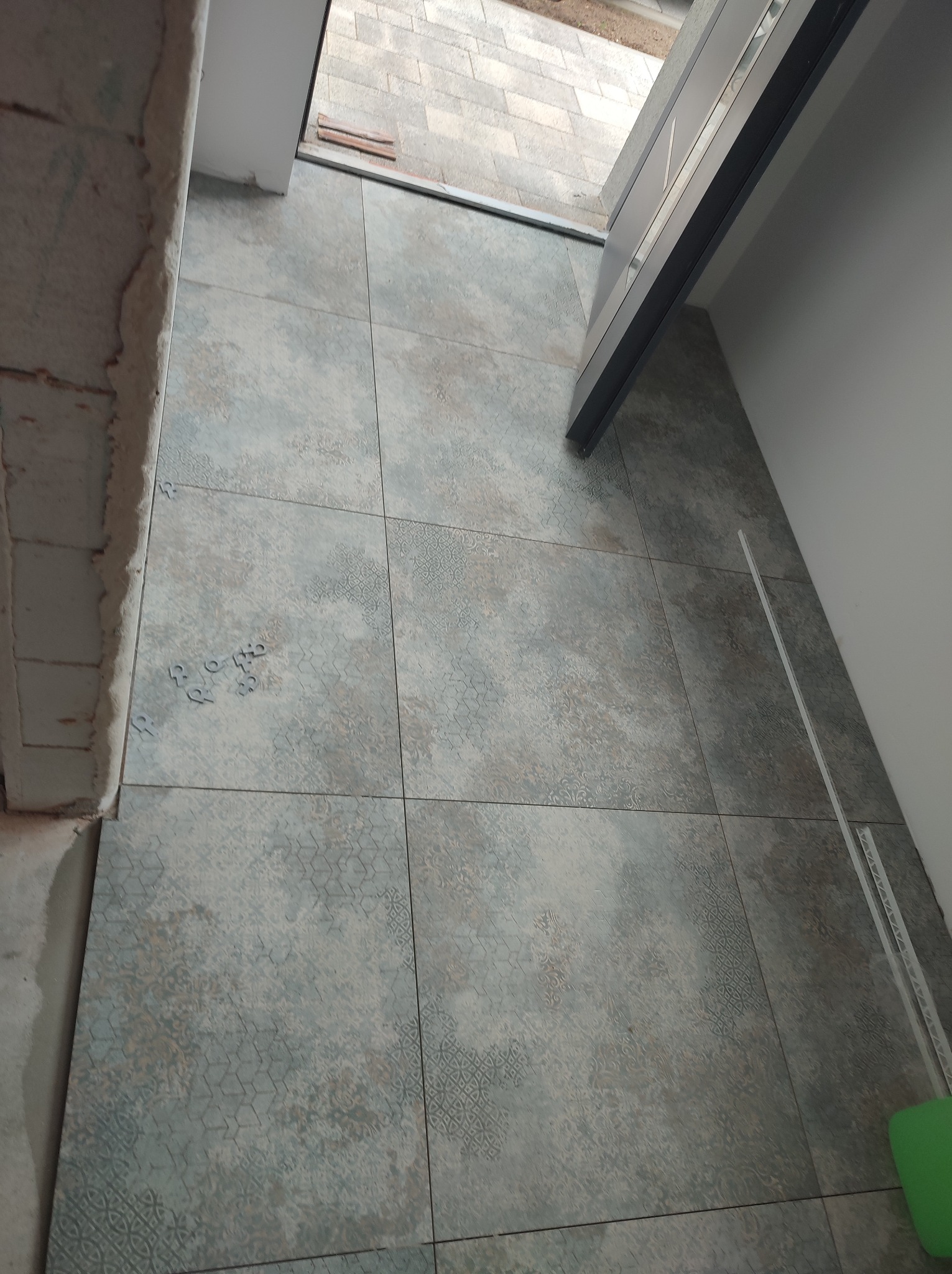 Ułożona podłoga z płytek w odcieniach szarości i beżu, widoczne elementy montażowe i fragment progu drzwiowego. Płytki z delikatnym wzorem, świeżo położone.