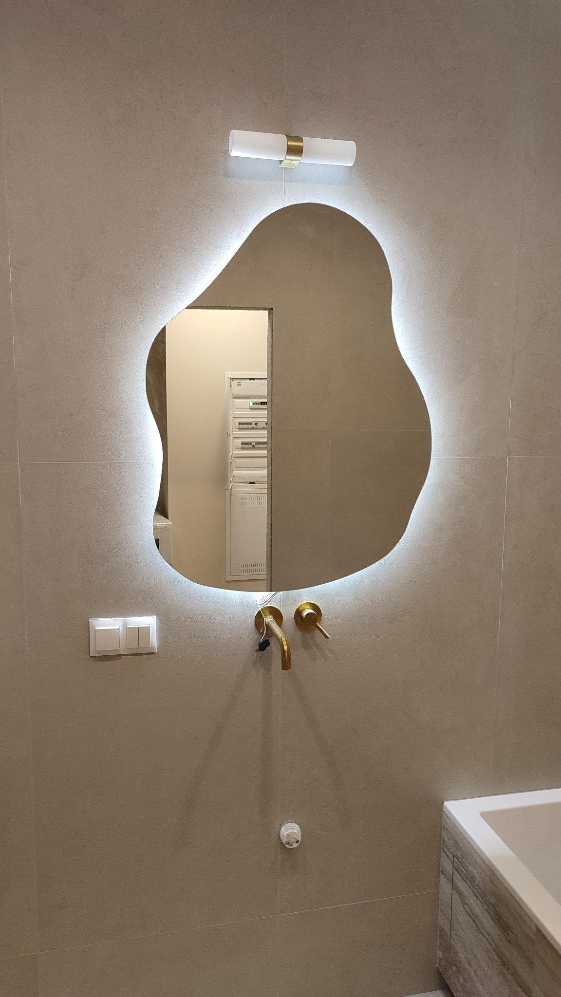 Beżowa łazienka z podświetlanym, asymetrycznym lustrem, złotą armaturą i płytkami imitującymi kamień. Widoczna szafka pod umywalką i biały włącznik światła.