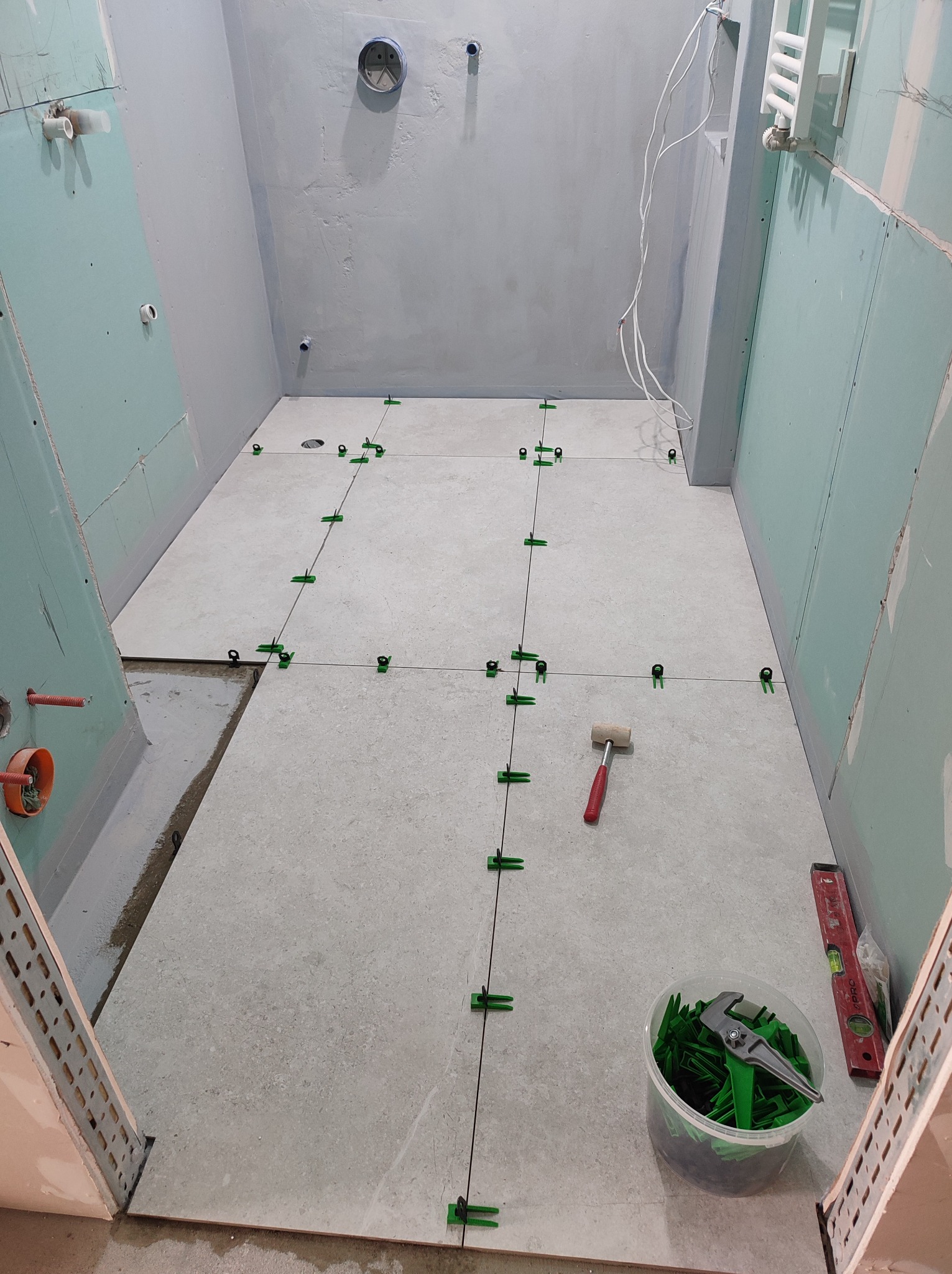 Układanie jasnych płytek podłogowych w łazience, z użyciem systemu poziomowania (klipsy i kliny). Widoczny młotek gumowy i poziomica. Ściany przygotowane pod wykończenie.