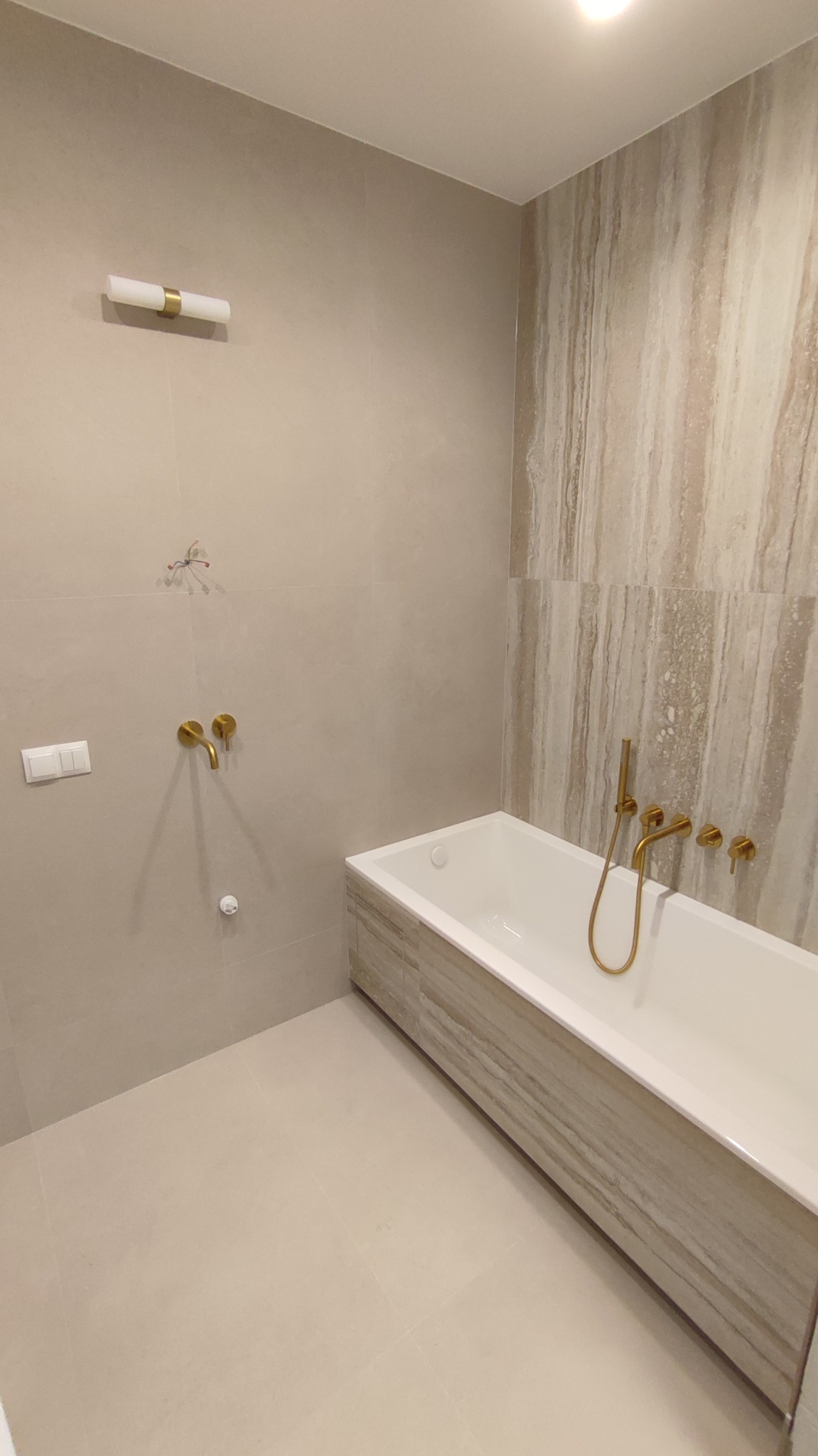 Jasna łazienka z beżowymi płytkami imitującymi marmur, białą wanną obudowaną płytkami i złotą armaturą. Widoczne włączniki i lampa nad wanną.