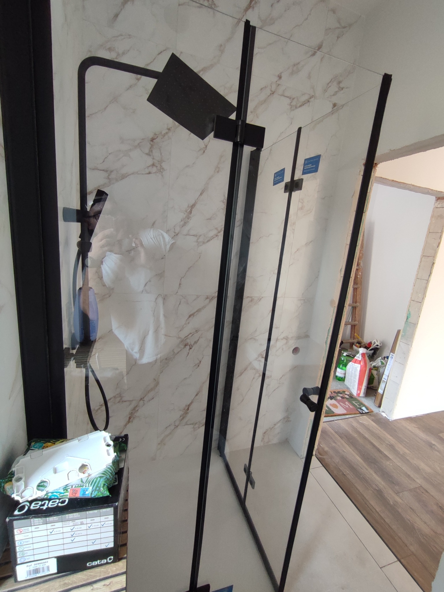 Nowoczesna kabina prysznicowa z czarnymi profilami i deszczownicą, marmurowe płytki na ścianie, widoczne akcesoria montażowe. Minimalistyczny design.