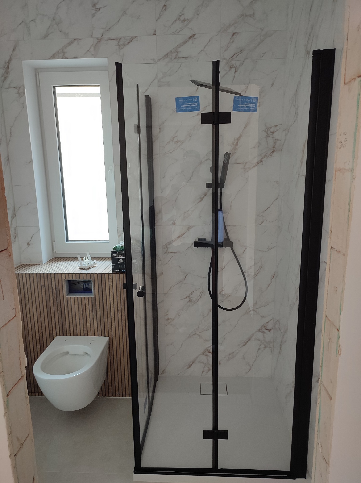 Nowoczesna kabina prysznicowa z czarnymi profilami i deszczownicą, marmurowe płytki na ścianach, podwieszana toaleta i drewniane panele w wykończonej łazience.