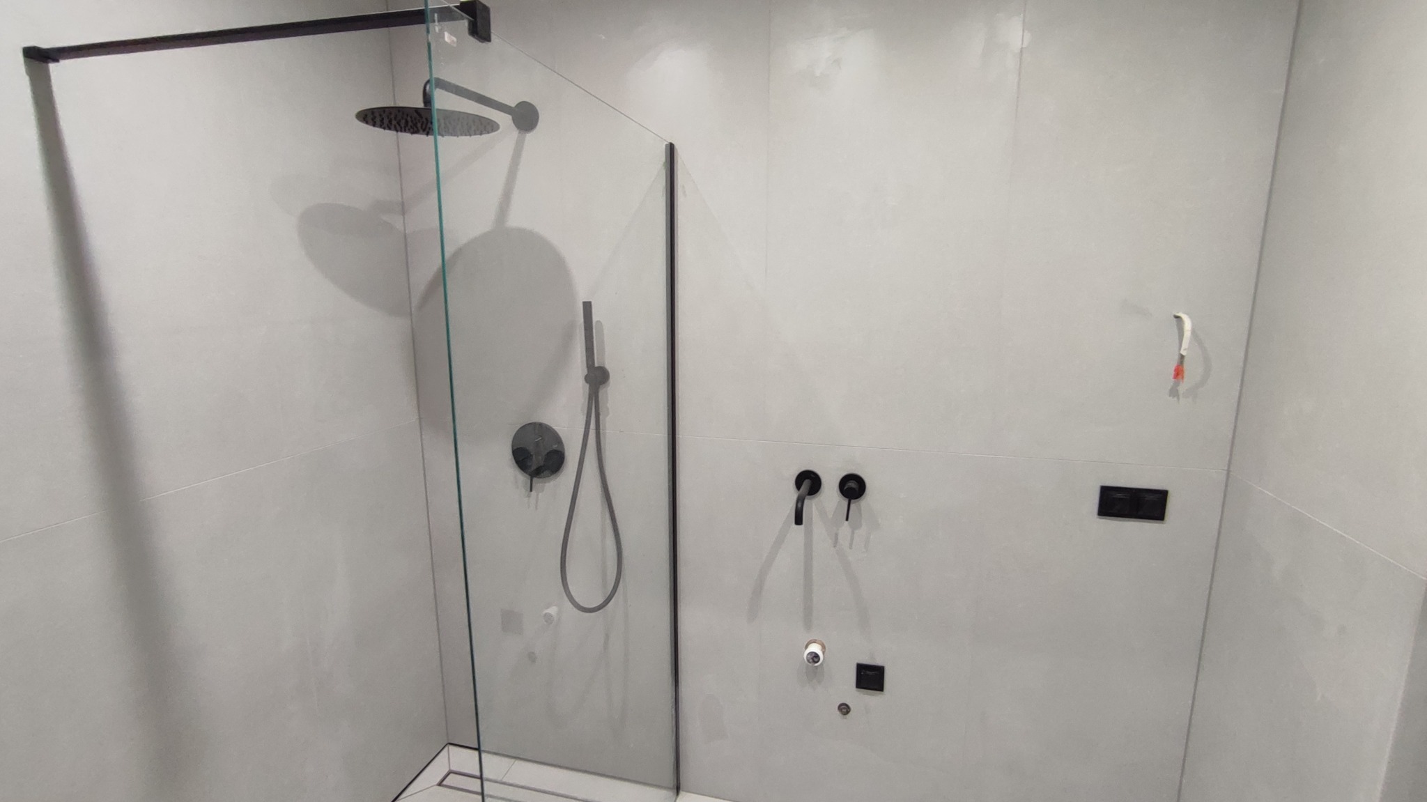 Nowoczesna kabina prysznicowa walk-in z czarną armaturą na tle jasnych, gładkich płytek. Minimalistyczny design, widoczne detale wykończenia.