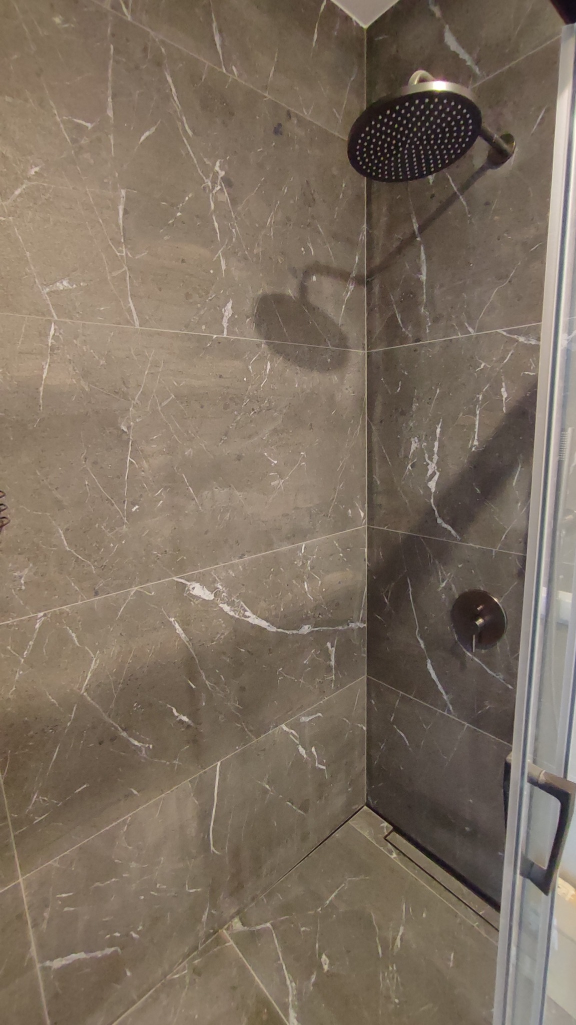 Kabina prysznicowa z szarymi płytkami imitującymi marmur, deszczownica i szklane drzwi. Widoczny fragment odpływu liniowego.