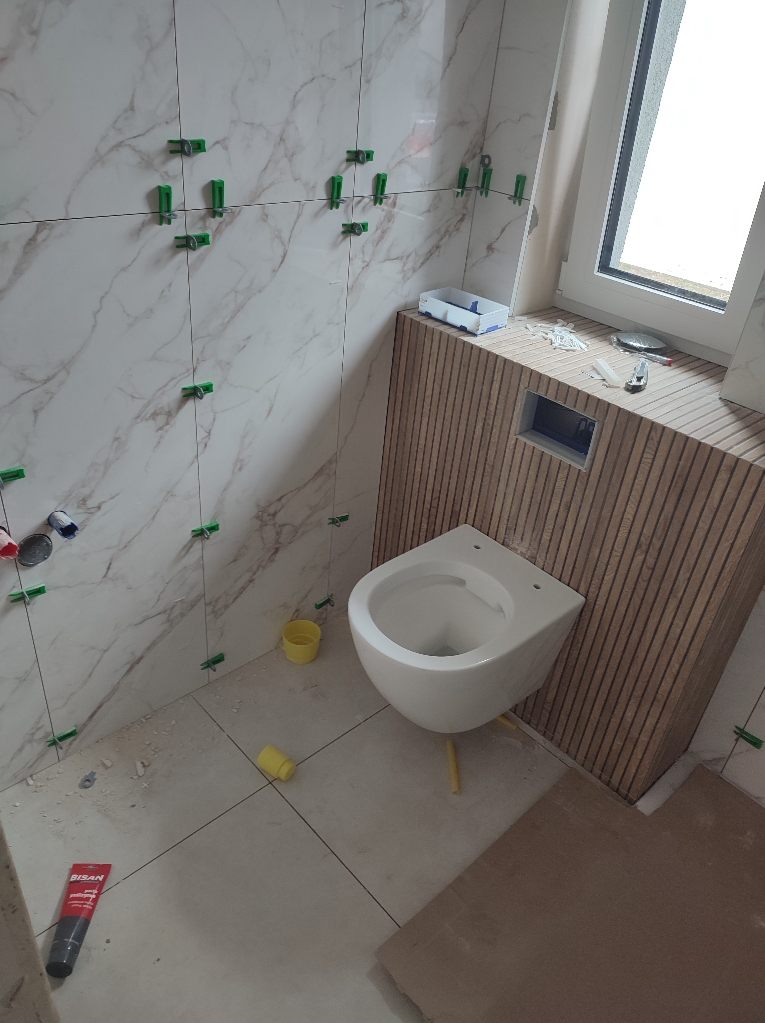 Wnętrze łazienki w trakcie prac wykończeniowych: białe płytki imitujące marmur z zielonymi krzyżykami, podwieszana toaleta na tle drewnianej ściany z desek.