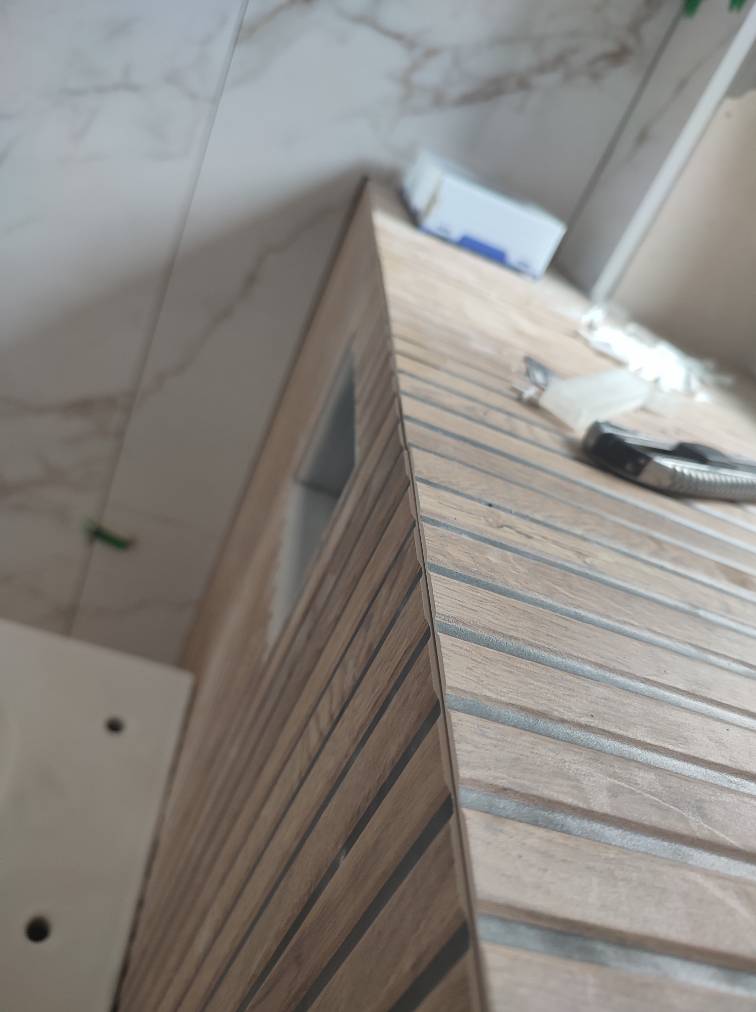 Ukończona obudowa z drewnopodobnych paneli ściennych w łazience, z widocznym narożnikiem i fragmentem umywalki. Na panelach leżą narzędzia.