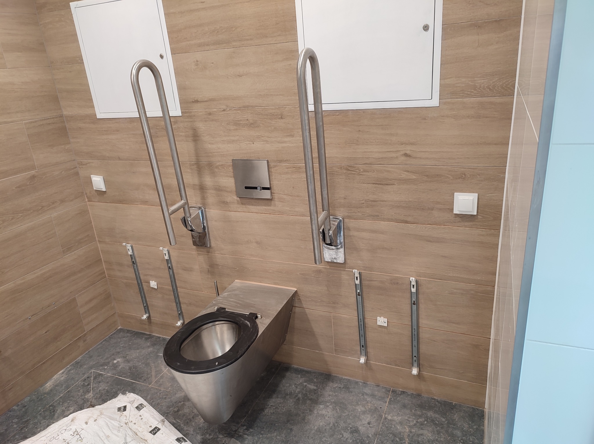 Nowoczesna toaleta podwieszana ze stali nierdzewnej, z uchwytami dla niepełnosprawnych na tle płytek imitujących drewno. Widoczne elementy montażowe.