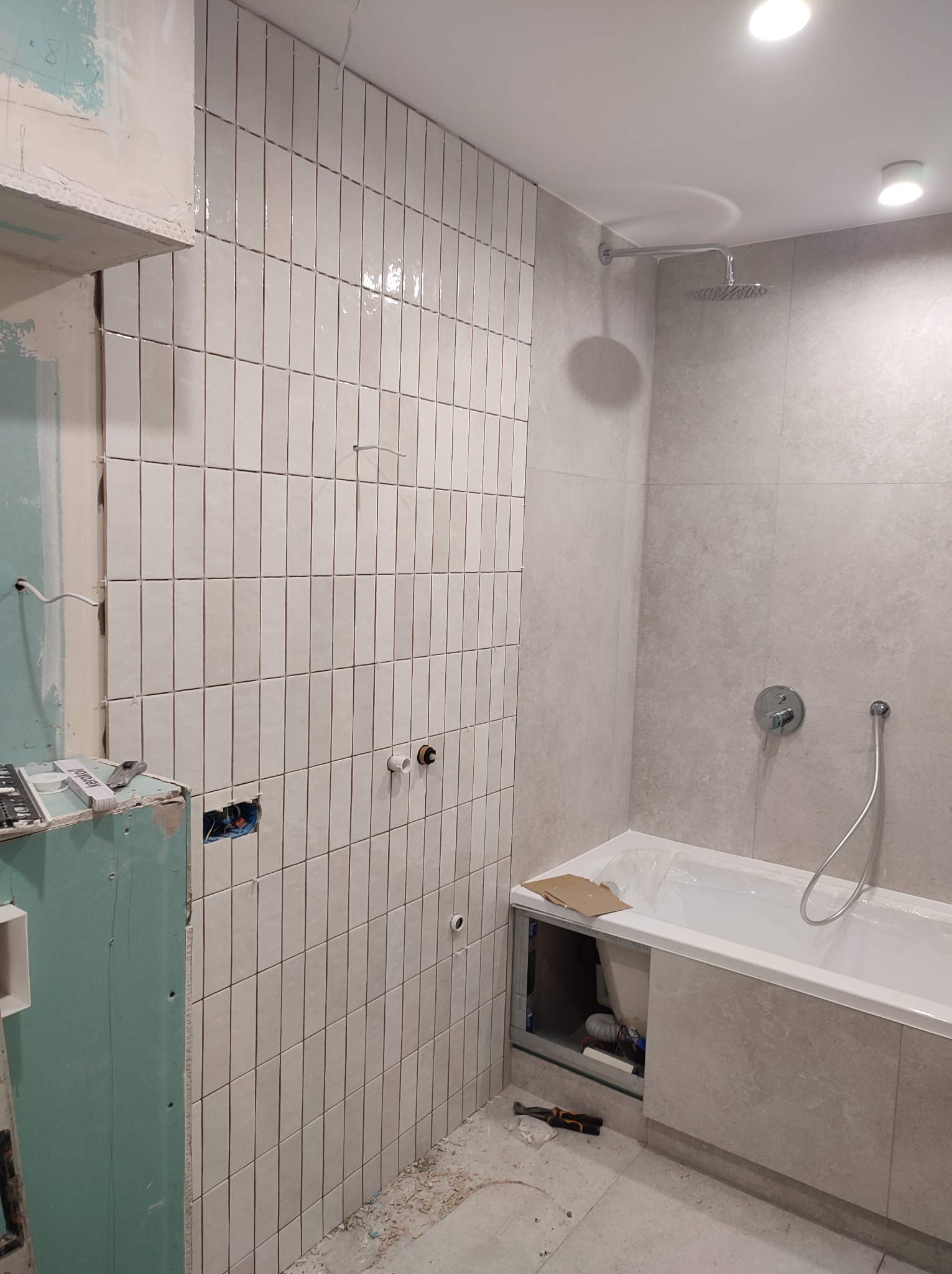 Białe, błyszczące, pionowo ułożone płytki na ścianie łazienki w trakcie montażu. Widoczne narzędzia, fragment wanny i instalacja prysznicowa. Surowy stan wykończenia.