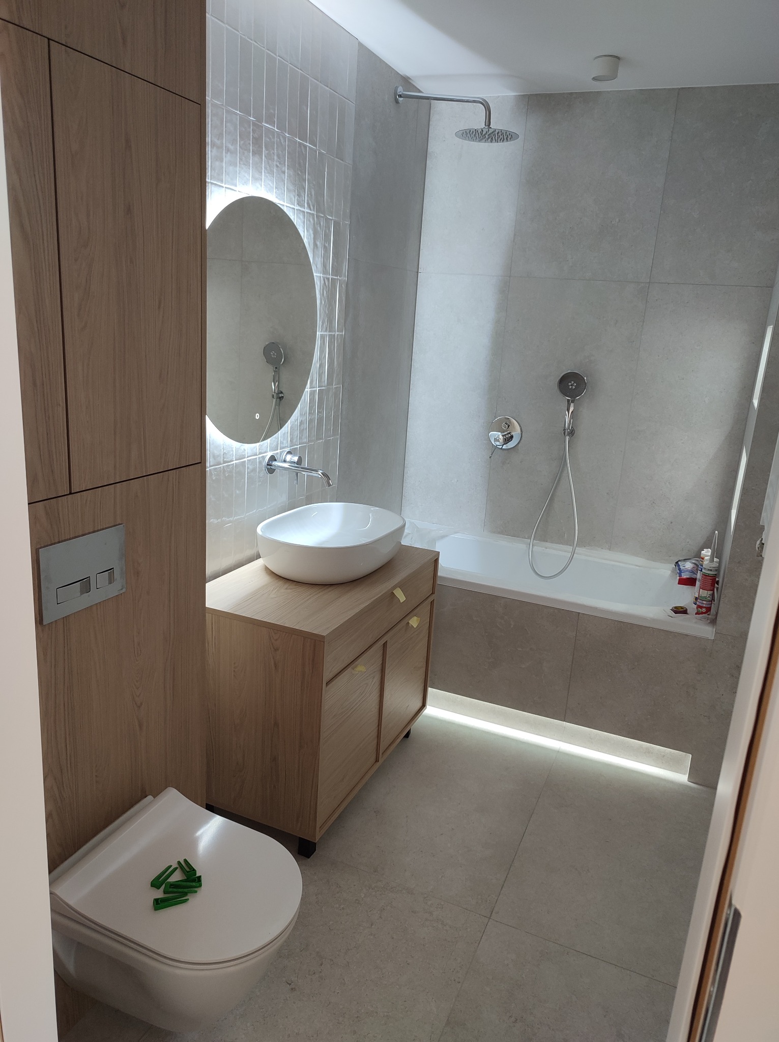 Jasna łazienka po remoncie: owalne lustro z podświetleniem, umywalka na szafce z drewnianym fornirem, podwieszana toaleta z zielonymi zaślepkami, wanna z prysznicem.