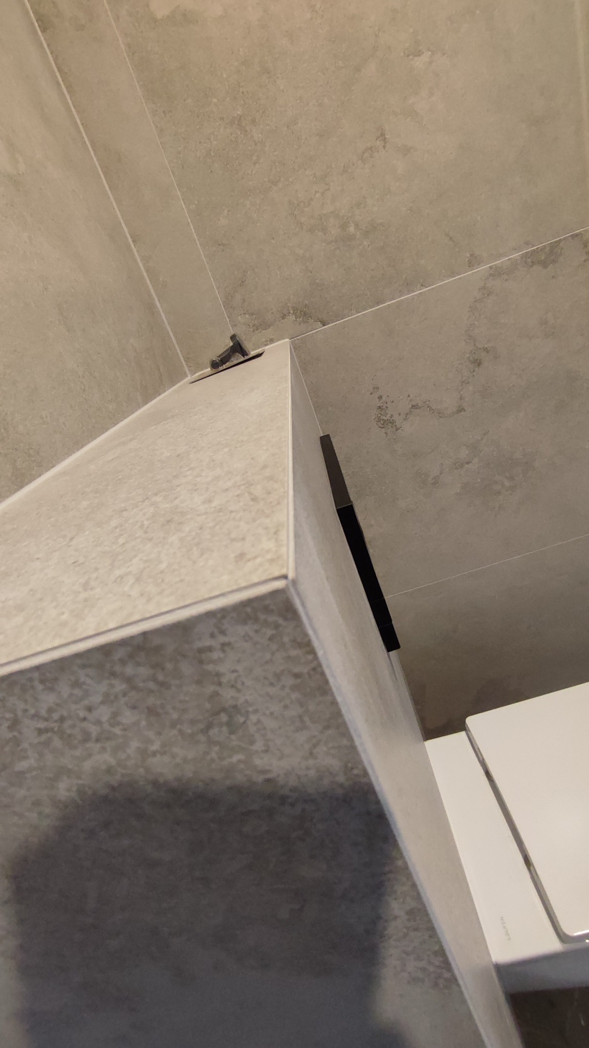 Wykończona obudowa WC z szarych płytek w łazience. Widoczny fragment toalety i ściana z identycznymi płytkami. Użyto metalowego profilu wykończeniowego.