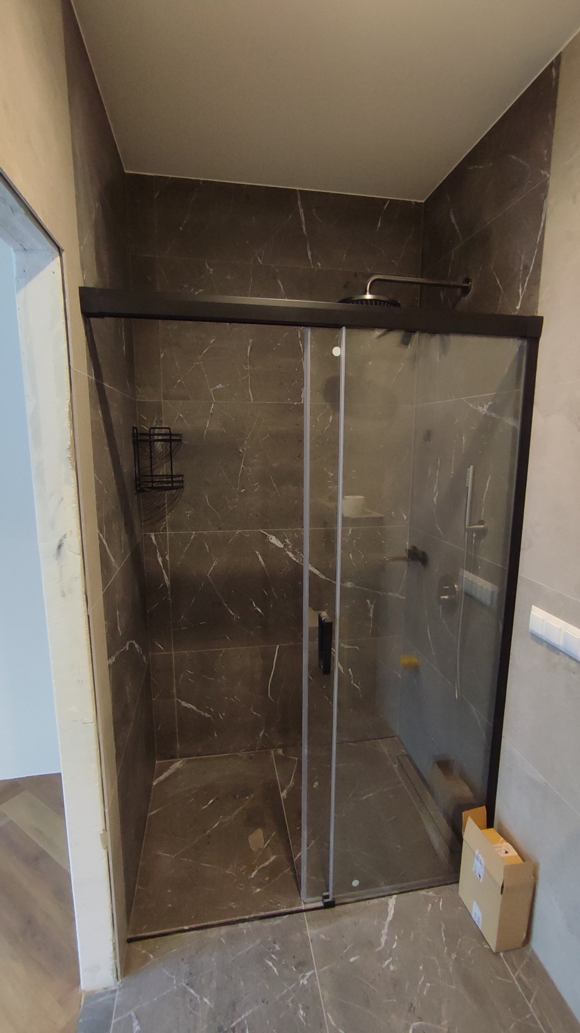 Nowoczesna kabina prysznicowa z przesuwnymi drzwiami, wykończona płytkami imitującymi marmur. Widoczny czarny profil kabiny i kartonowe pudełko w rogu.