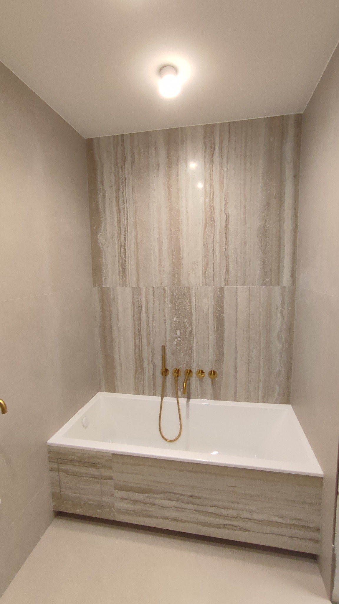 Minimalistyczna łazienka z wanną obudowaną płytkami imitującymi trawertyn, złota armatura, oświetlenie sufitowe. Neutralna kolorystyka, nowoczesny design.