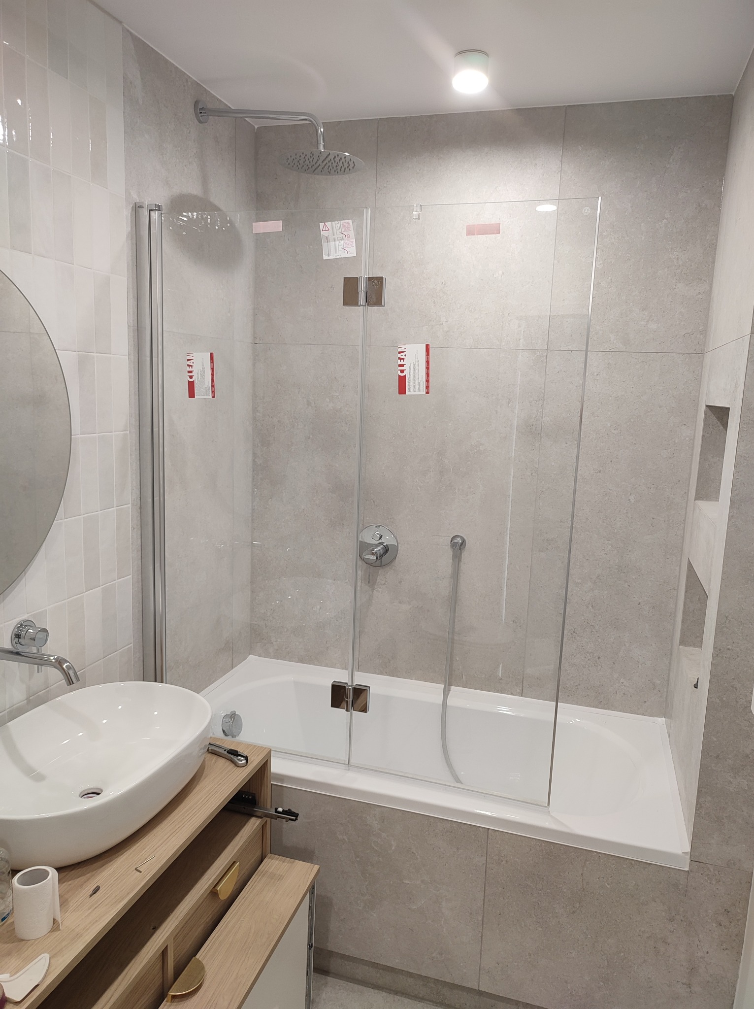 Nowoczesna łazienka w szarościach z owalną umywalką na drewnianej szafce i kabiną prysznicową z transparentnego szkła. Minimalistyczny design, subtelne oświetlenie.