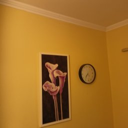 Usługi remotowo-budowlane Marcin Brwinowski - Żółta ściana z obrazem kalii i zegarem. Biały sufit z listwą. Ciepłe oświetlenie. Ujęcie z dołu, widoczny fragment rogu pokoju.