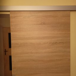 Usługi remotowo-budowlane Marcin Brwinowski - Zamknięte, przesuwne drzwi z drewnianym wzorem, zamontowane na metalowej szynie. W tle widoczne fragmenty ściany i drzwi wejściowych.