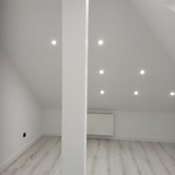 Usługi remotowo-budowlane Marcin Brwinowski - Jasne poddasze po remoncie: białe ściany, drewnopodobna podłoga, kolumna, oświetlenie punktowe w suficie skośnym. Widoczny grzejnik i gniazdko elektryczne.