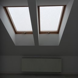Usługi remotowo-budowlane Marcin Brwinowski - Poddasze z dwoma oknami dachowymi w drewnianych ramach, białe ściany i grzejnik. Minimalistyczne wykończenie wnętrza, jasna podłoga. Ujęcie od dołu.