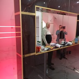 Usługi remotowo-budowlane Marcin Brwinowski - Wykończona łazienka z czarnymi płytkami imitującymi marmur, złotymi ramami i podświetleniem LED. W lustrze odbija się osoba robiąca zdjęcie.