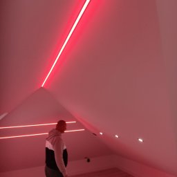 Usługi remotowo-budowlane Marcin Brwinowski - Podświetlone, czerwone listwy LED w skosach poddasza, tworzące nowoczesny efekt oświetleniowy. Widoczna osoba w pomieszczeniu, podkreślająca skalę i charakter wnętrza.