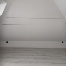 Usługi remotowo-budowlane Marcin Brwinowski - Wnętrze poddasza po remoncie we Wszeborach: białe ściany, jasna podłoga, widoczne listwy i gniazdka elektryczne. Skos dachu i minimalistyczny styl.