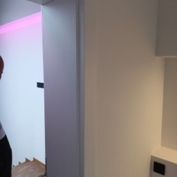Usługi remotowo-budowlane Marcin Brwinowski - Nowoczesne wnętrze po remoncie: białe ściany, minimalistyczne drzwi z czarnymi zawiasami, widoczny fragment schodów i oświetlenie LED w kolorze różowym.