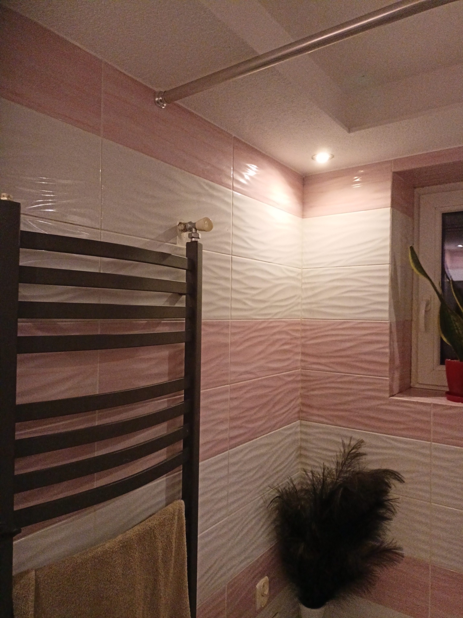Wykończona łazienka z grzejnikiem drabinkowym, ręcznikiem i dekoracyjnymi płytkami w odcieniach różu i bieli. Widoczne oświetlenie sufitowe i roślina w doniczce.