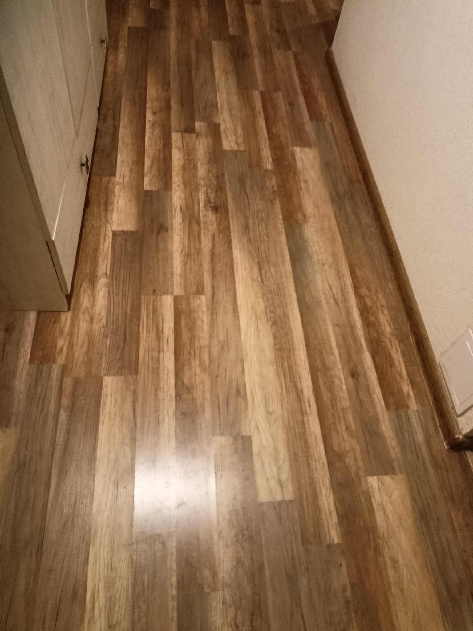 Ułożona podłoga z paneli imitujących drewno w odcieniach brązu, widok z góry, fragment szafki i ściany w tle. Układ paneli w jodełkę.