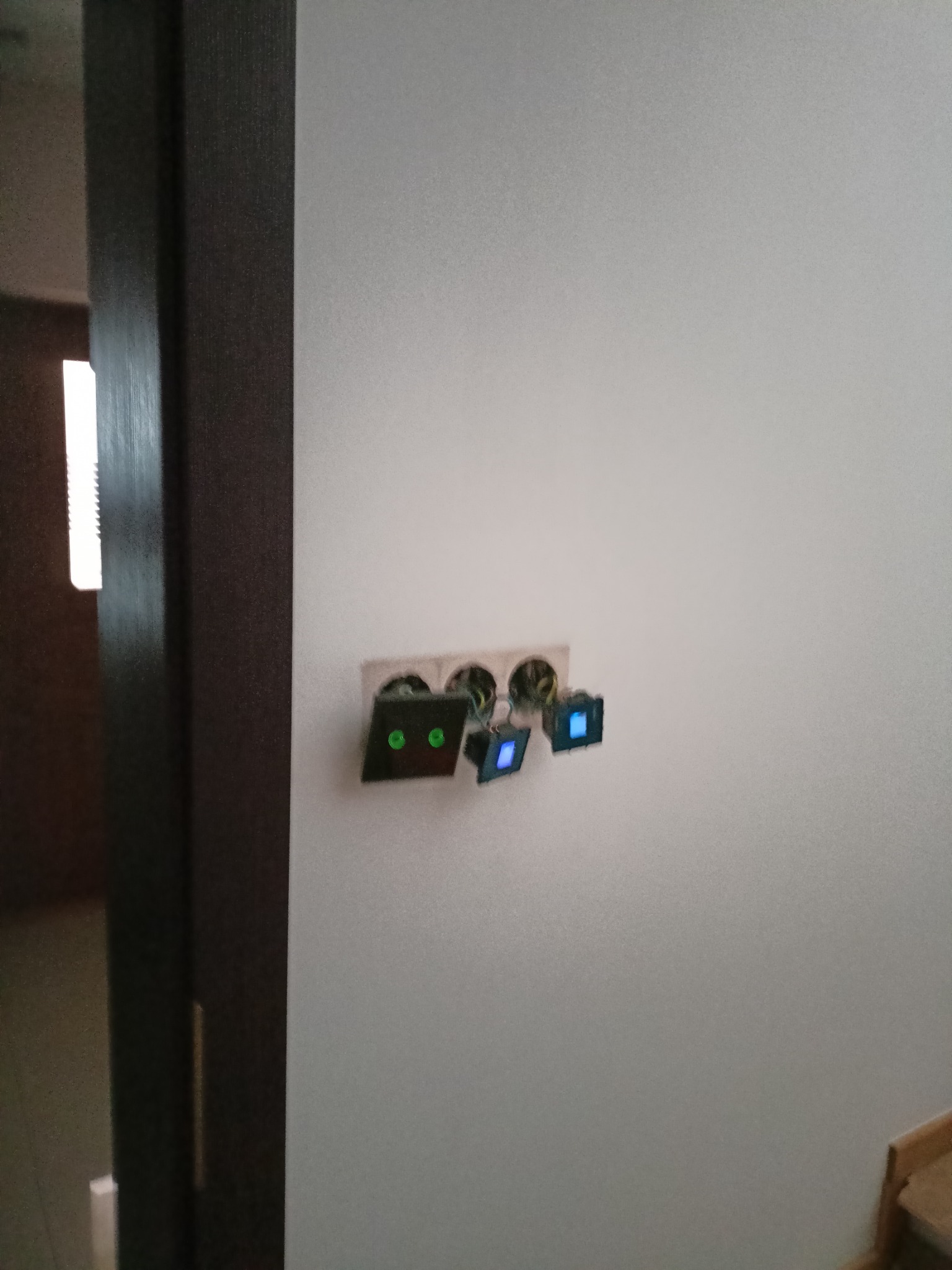 Biała ściana z zamontowanymi, nowoczesnymi gniazdkami elektrycznymi z podświetleniem LED w kolorach zielonym i niebieskim. Widoczny fragment ościeżnicy drzwi.