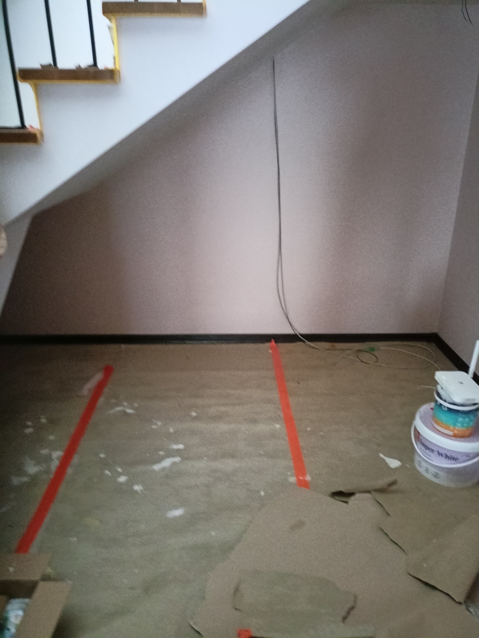 Pokój w trakcie przygotowań do malowania: zabezpieczona podłoga folią i taśmą, widoczne puszki z farbą, ściany w jasnym odcieniu, schody w tle. Przewody zwisają ze ściany.