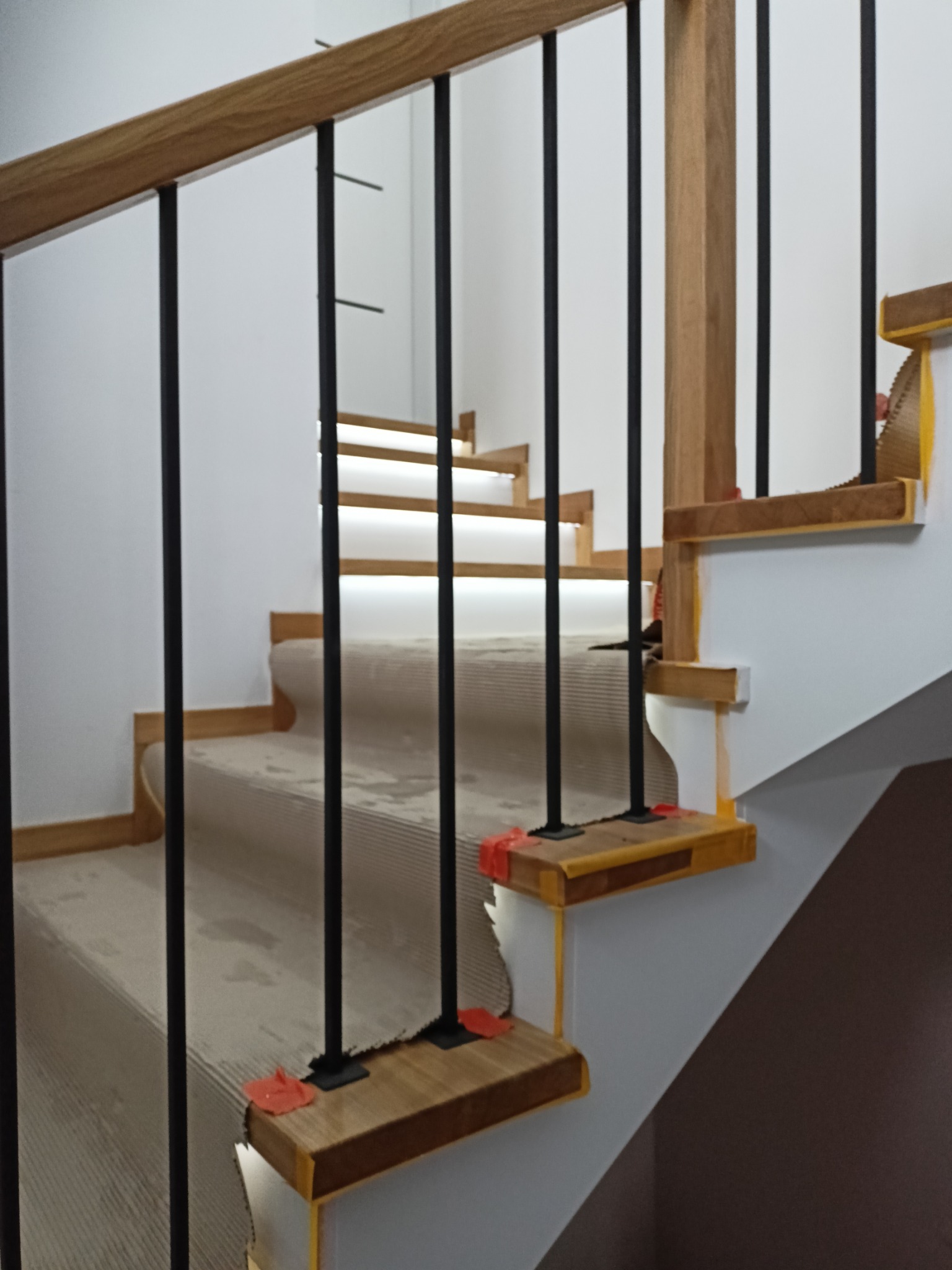 Schody drewniane z czarną balustradą i podświetleniem LED, wykończone wykładziną. Detale zabezpieczone taśmą malarską, ukazujące etap prac wykończeniowych.