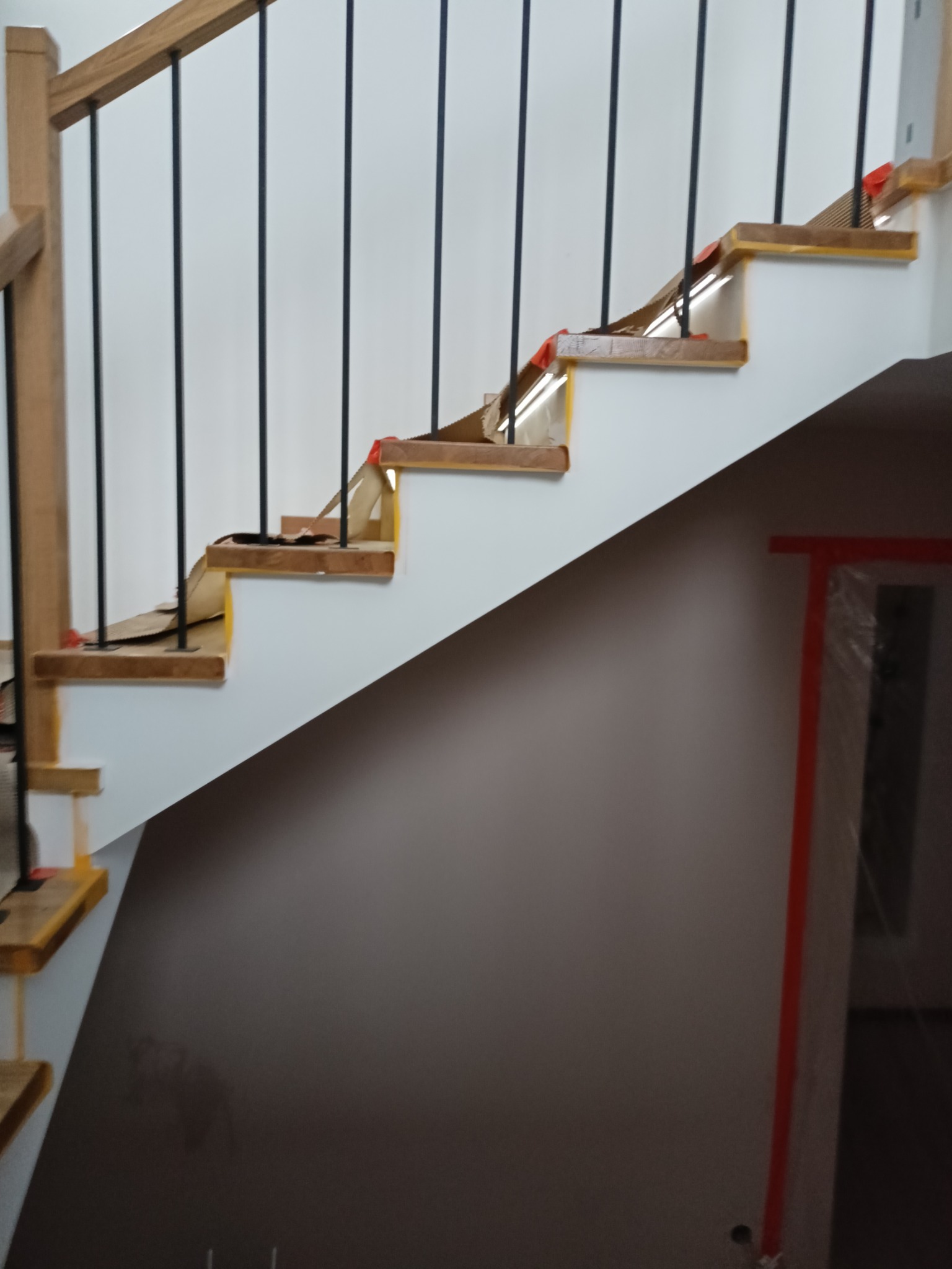 Schody z drewnianymi stopniami i czarną balustradą w trakcie zabezpieczania folią malarską. Ściany białe, widoczne przygotowania do prac wykończeniowych.