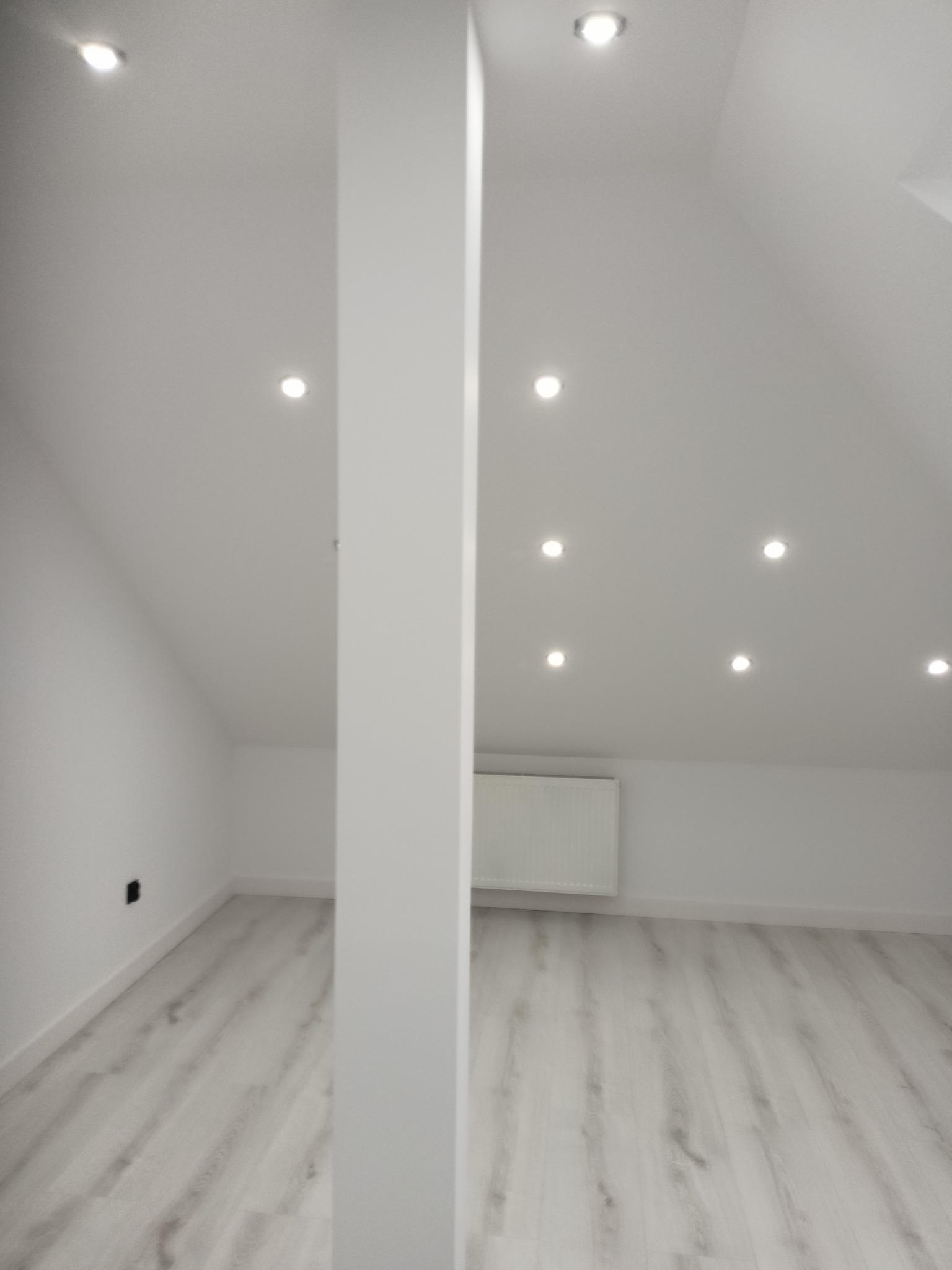 Jasne poddasze po remoncie: białe ściany, drewnopodobna podłoga, kolumna, oświetlenie punktowe w suficie skośnym. Widoczny grzejnik i gniazdko elektryczne.