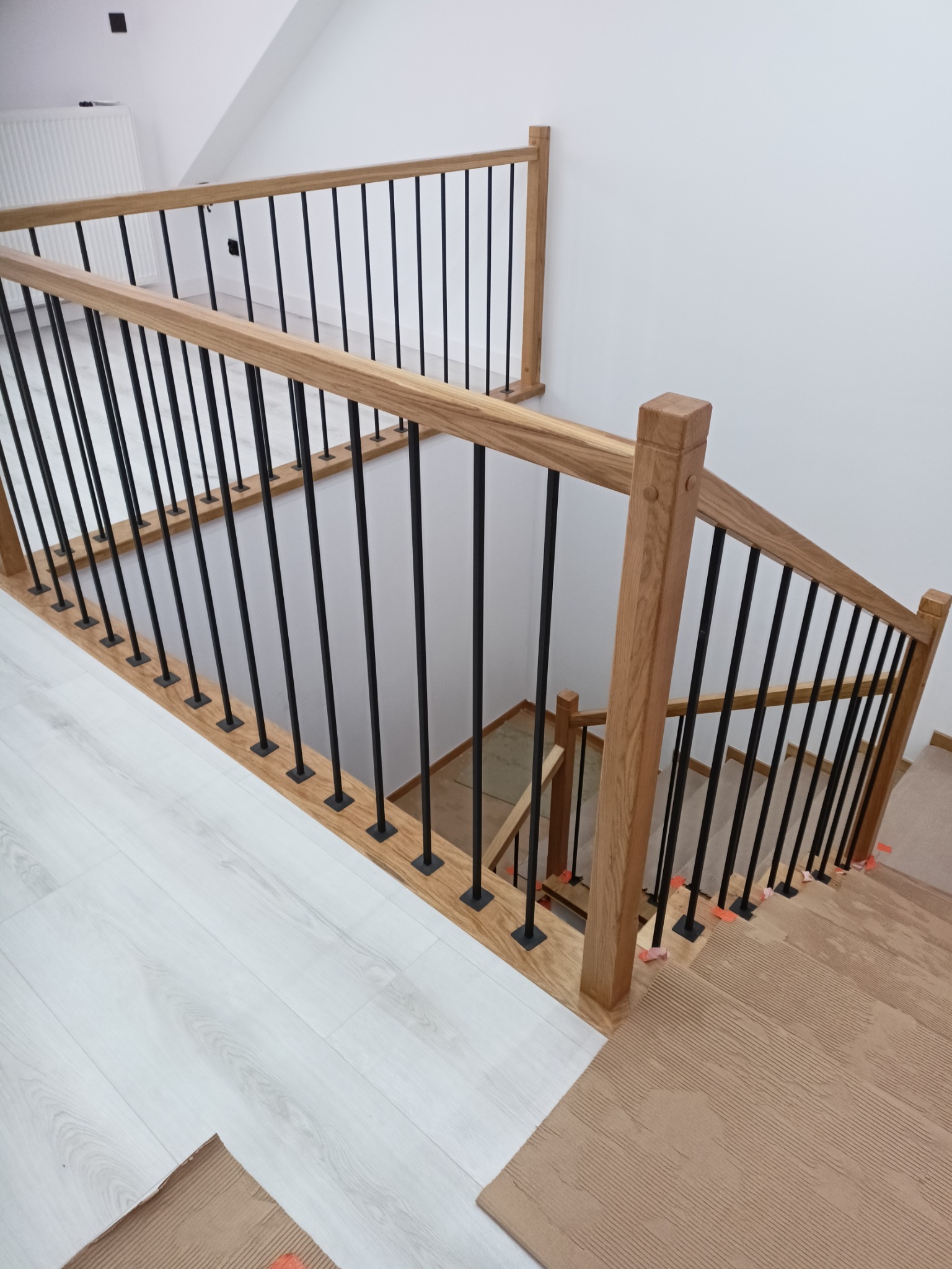 Nowoczesne schody dębowe z czarnymi balustradami, widok z góry. Jasne drewno kontrastuje z bielą ścian i podłogi. Detale wykończenia, kąt ujęcia podkreśla geometrię.