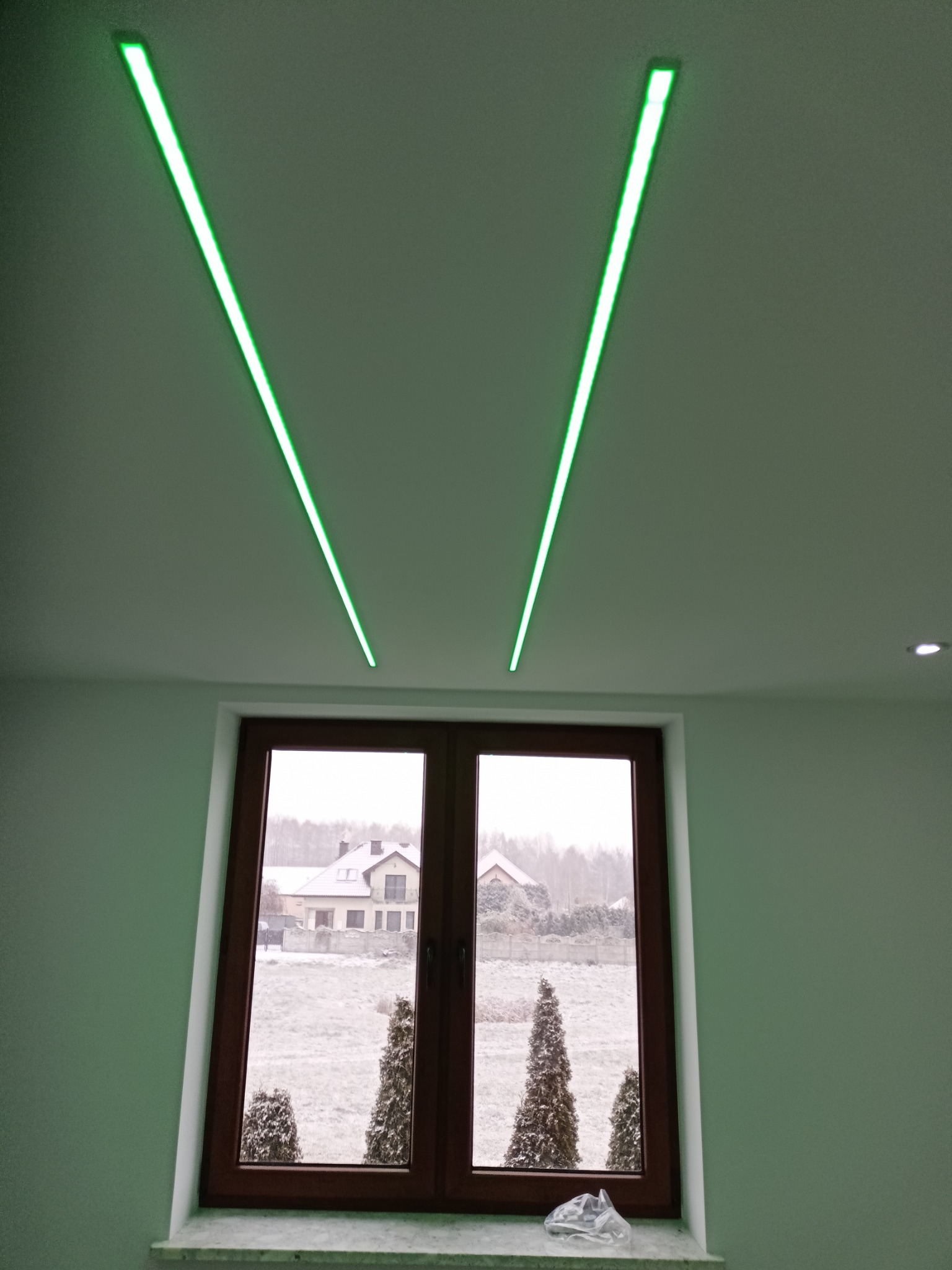 Nowoczesne oświetlenie sufitowe LED w kształcie litery V, okno z widokiem na zimowy krajobraz. Minimalistyczny design wnętrza z akcentem na zielone światło.