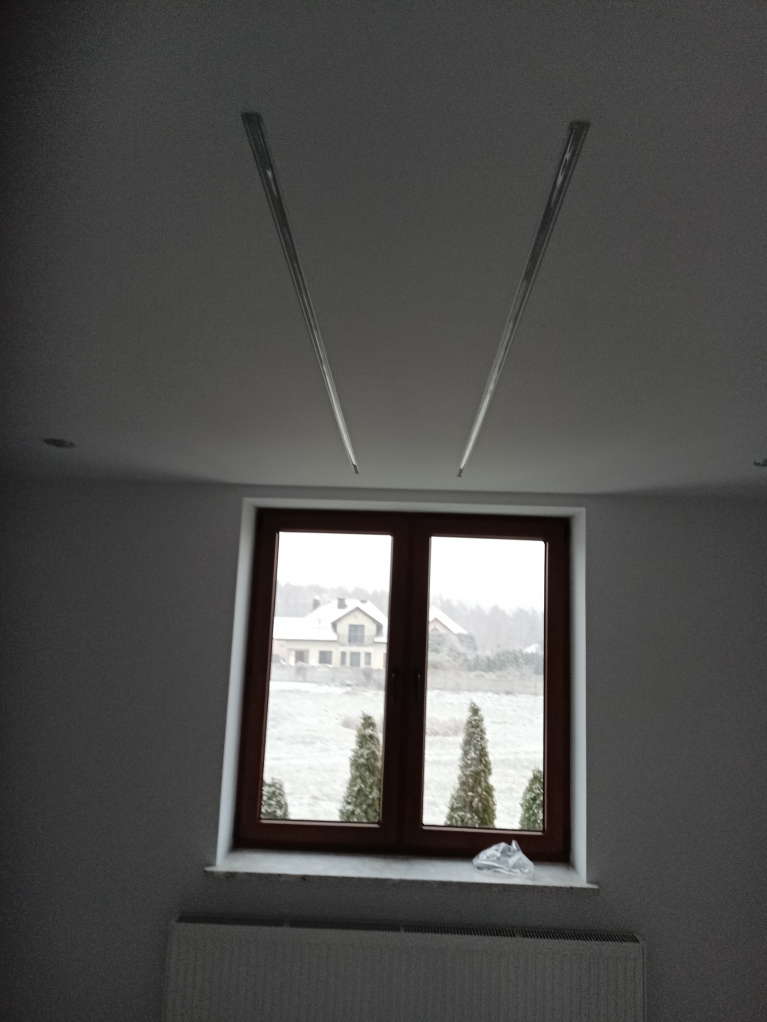 Wnętrze pokoju z oknem i grzejnikiem. Na suficie widoczne metalowe profile, prawdopodobnie przygotowane do montażu płyt kartonowo-gipsowych. Za oknem zimowy krajobraz.