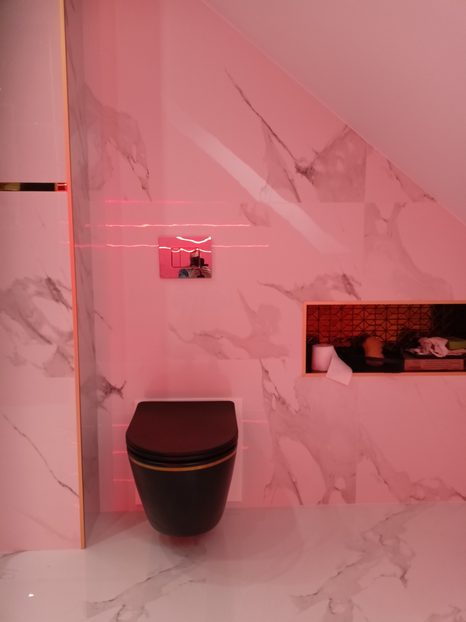 Nowoczesna toaleta w łazience z marmurowymi płytkami, podświetlona różowym światłem. Widoczna wnęka z dekoracyjną mozaiką i akcesoriami. Ujęcie z niskiego kąta.