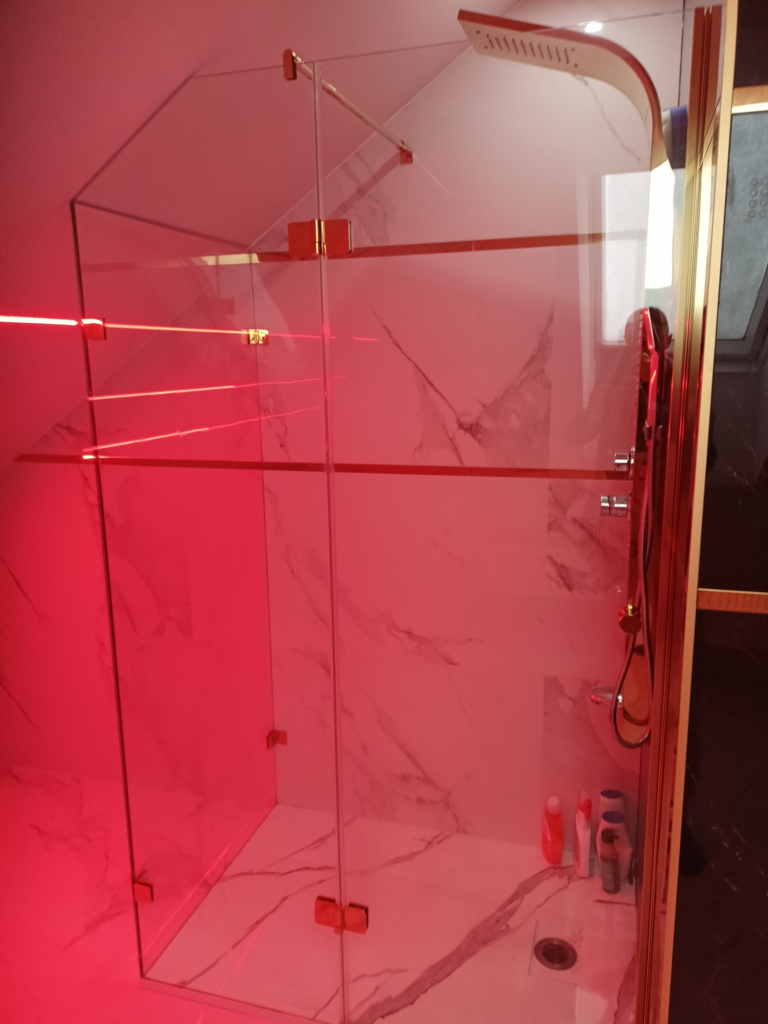 Nowoczesna kabina prysznicowa z marmurowymi ścianami, złotymi detalami i czerwonym oświetleniem LED. Widoczne szklane drzwi i deszczownica.