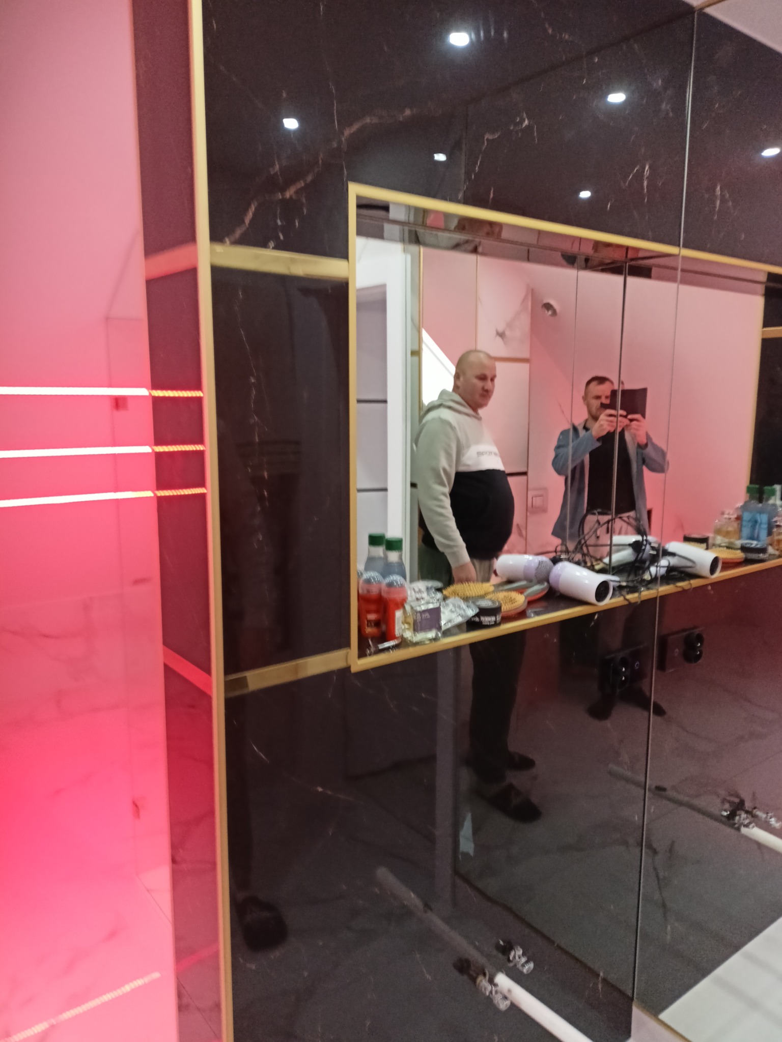 Wykończona łazienka z czarnymi płytkami imitującymi marmur, złotymi ramami i podświetleniem LED. W lustrze odbija się osoba robiąca zdjęcie.