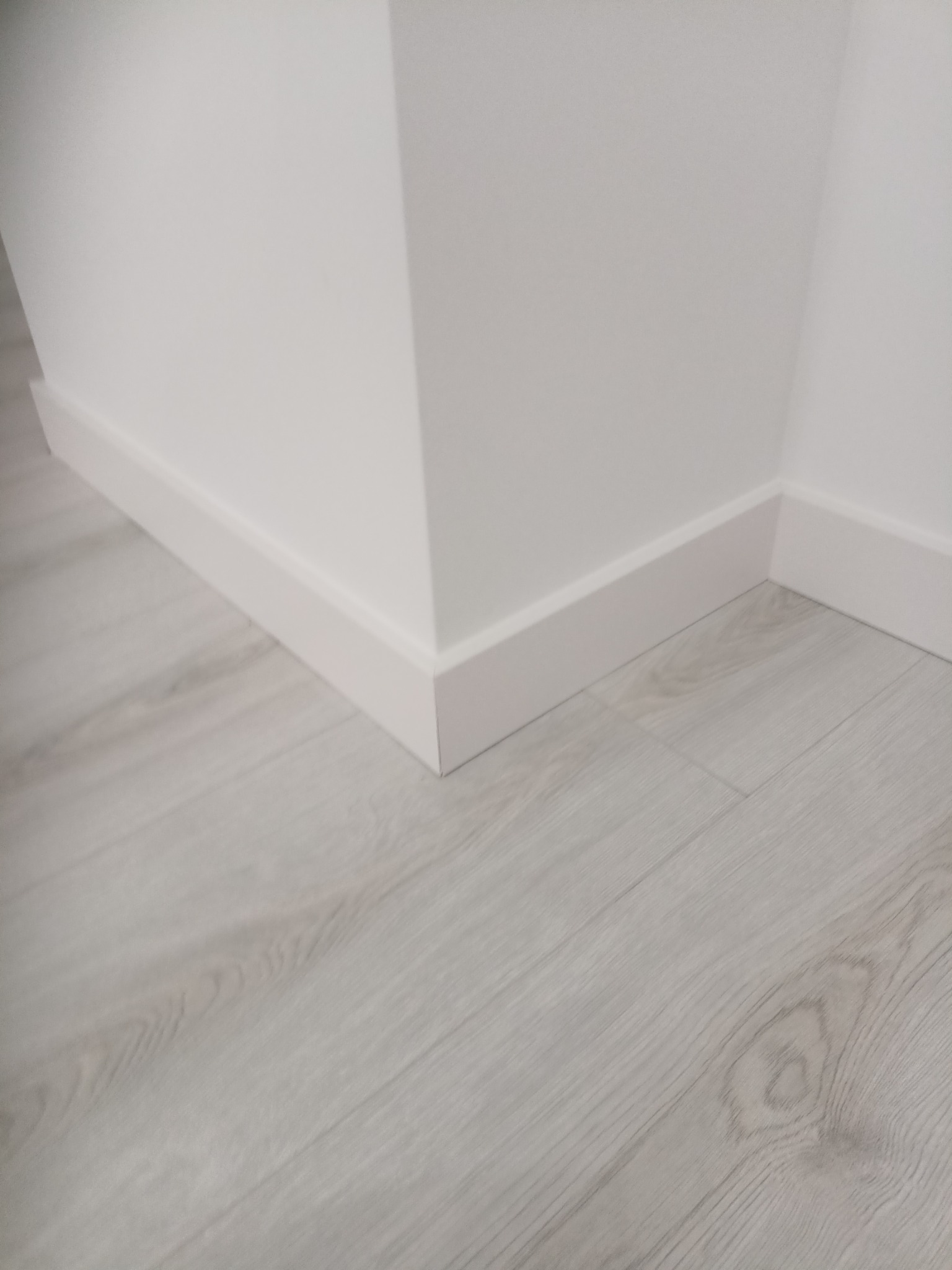 Białe listwy przypodłogowe zamontowane w rogu pomieszczenia, na podłodze panele w odcieniach szarości. Minimalistyczne wykończenie wnętrza, detale po remoncie.