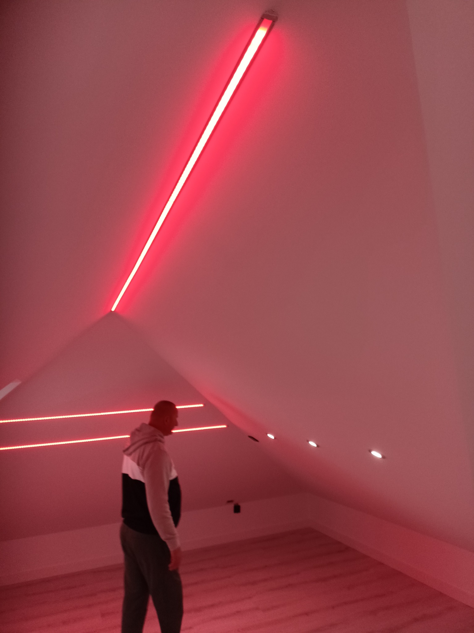 Podświetlone, czerwone listwy LED w skosach poddasza, tworzące nowoczesny efekt oświetleniowy. Widoczna osoba w pomieszczeniu, podkreślająca skalę i charakter wnętrza.