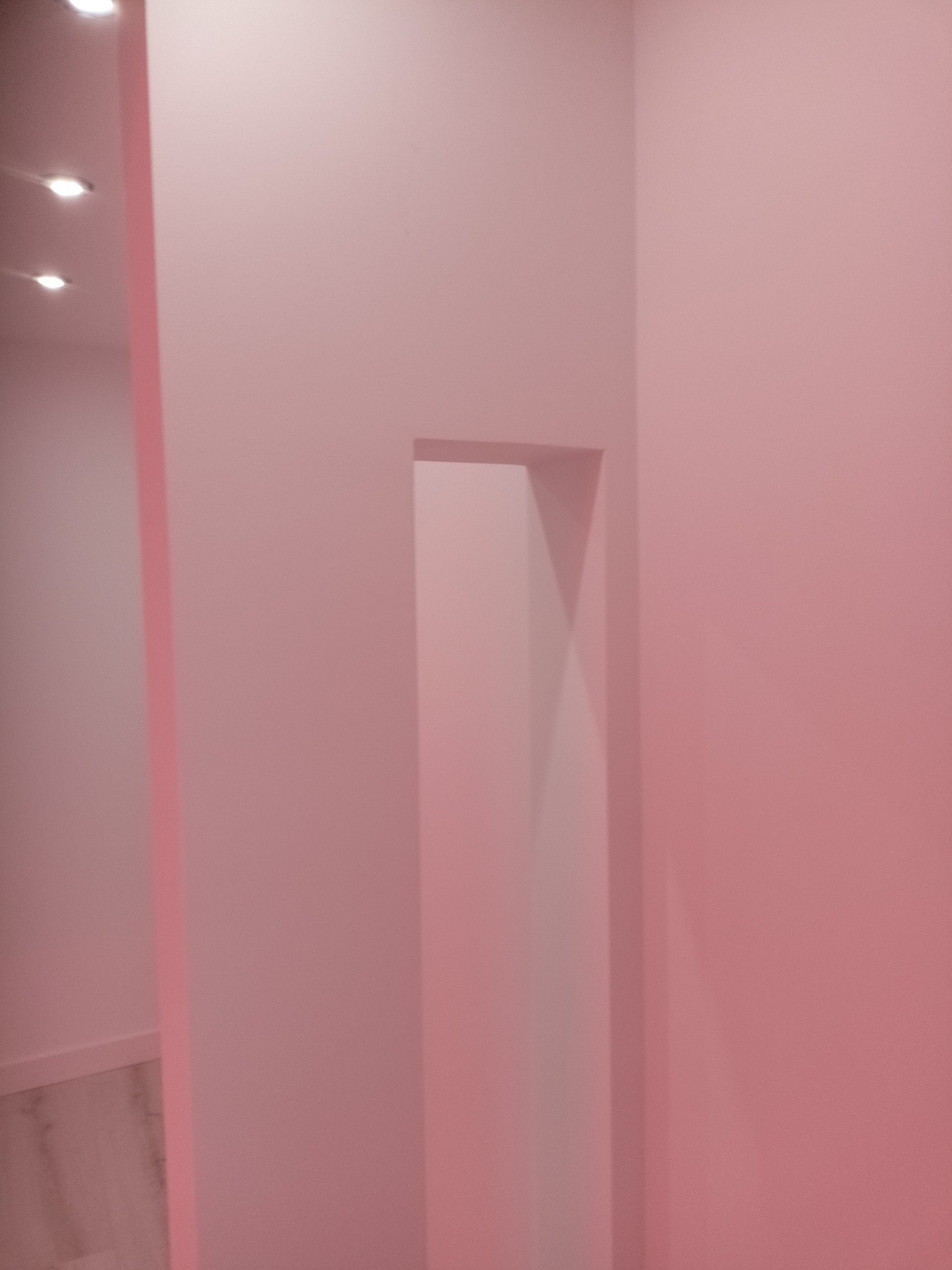 Pomalowane na różowo wnętrze z przejściem w ścianie. Widoczne oświetlenie sufitowe i fragment podłogi. Minimalistyczny design.